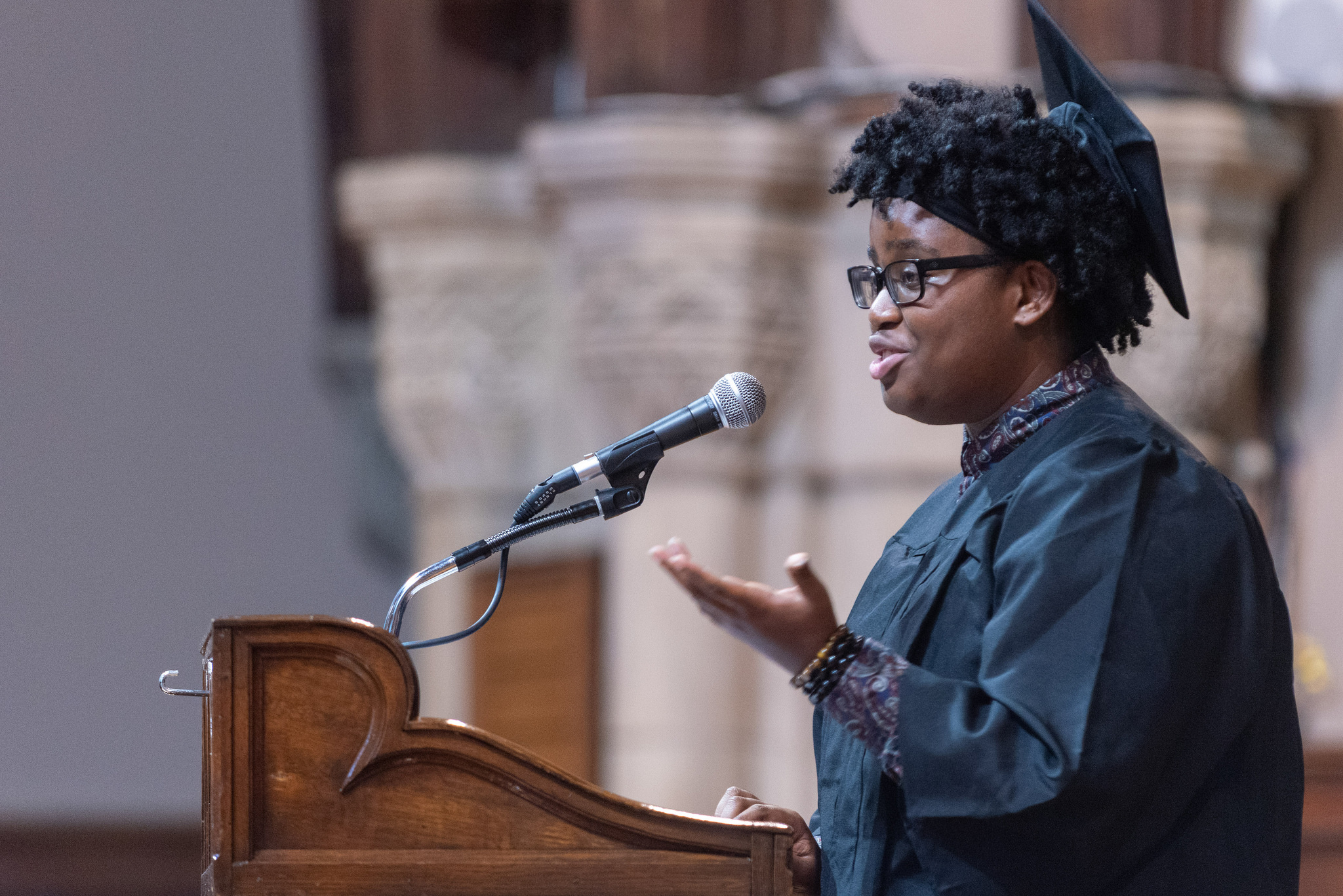 Tamar Ballard speaks in front of a podium. Ballard is wearing a dark robe and a dark cap.