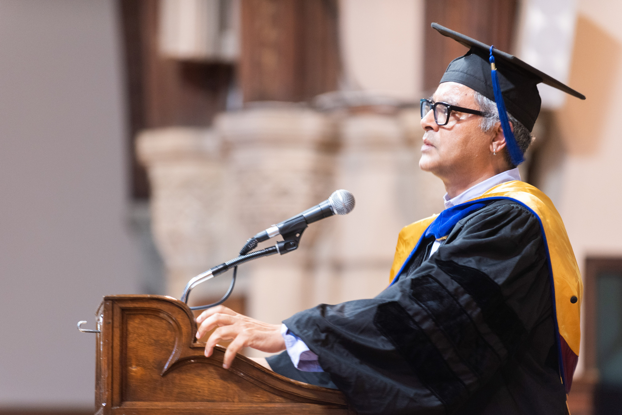 Amitava Kumar speaks in front of a podium. Kumar is wearing a black robe and black cap with a blue tassel.