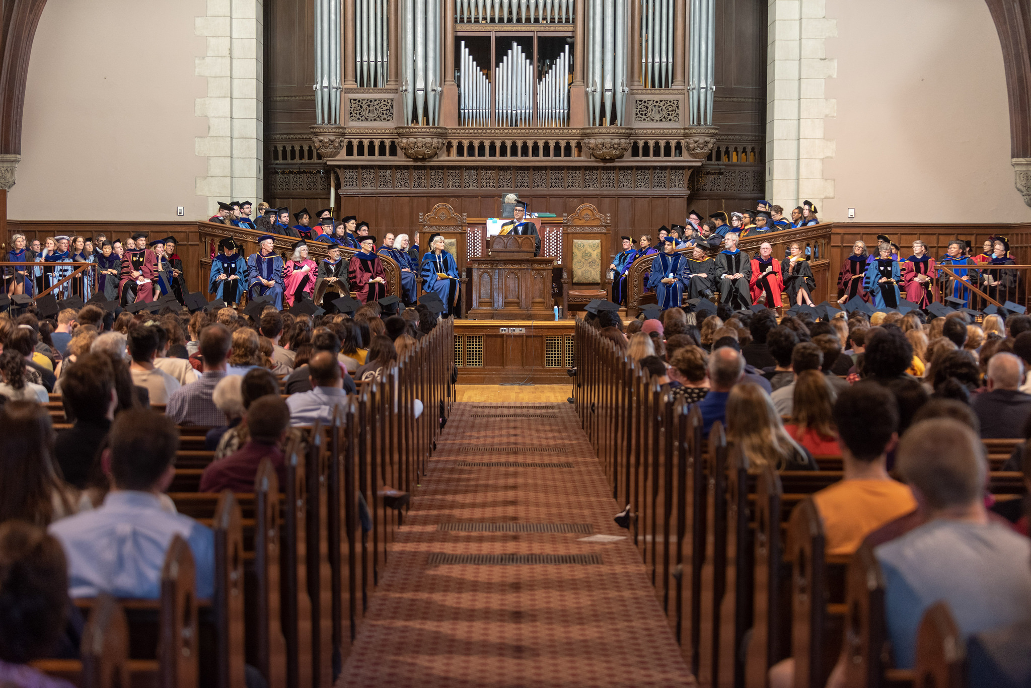 A view down the main aisle of the Chapel, with large crowds of people seated on either side. A man speaks in front of a podium in front of a giant church organ.