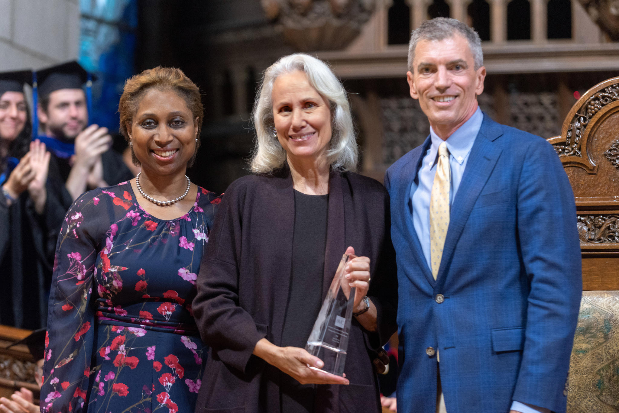 Three people stand in the chapel, smiling at the camera. One person holds a small glass award.