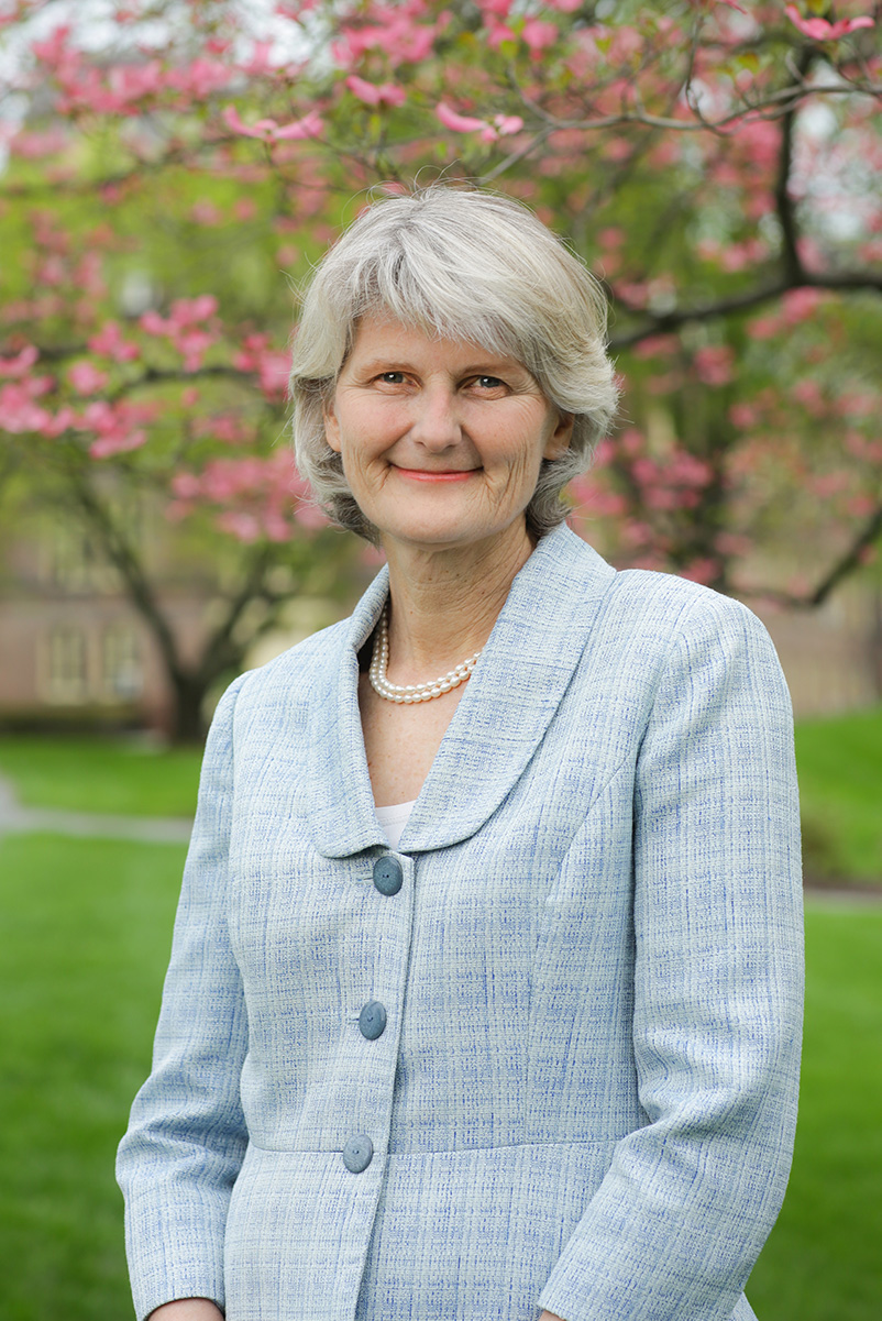 A person with mid-length gray hair and a light blue suit jacket smiles at the viewer. The person has a pearl necklace, and is standing outside, under a tree with pink blossoms.