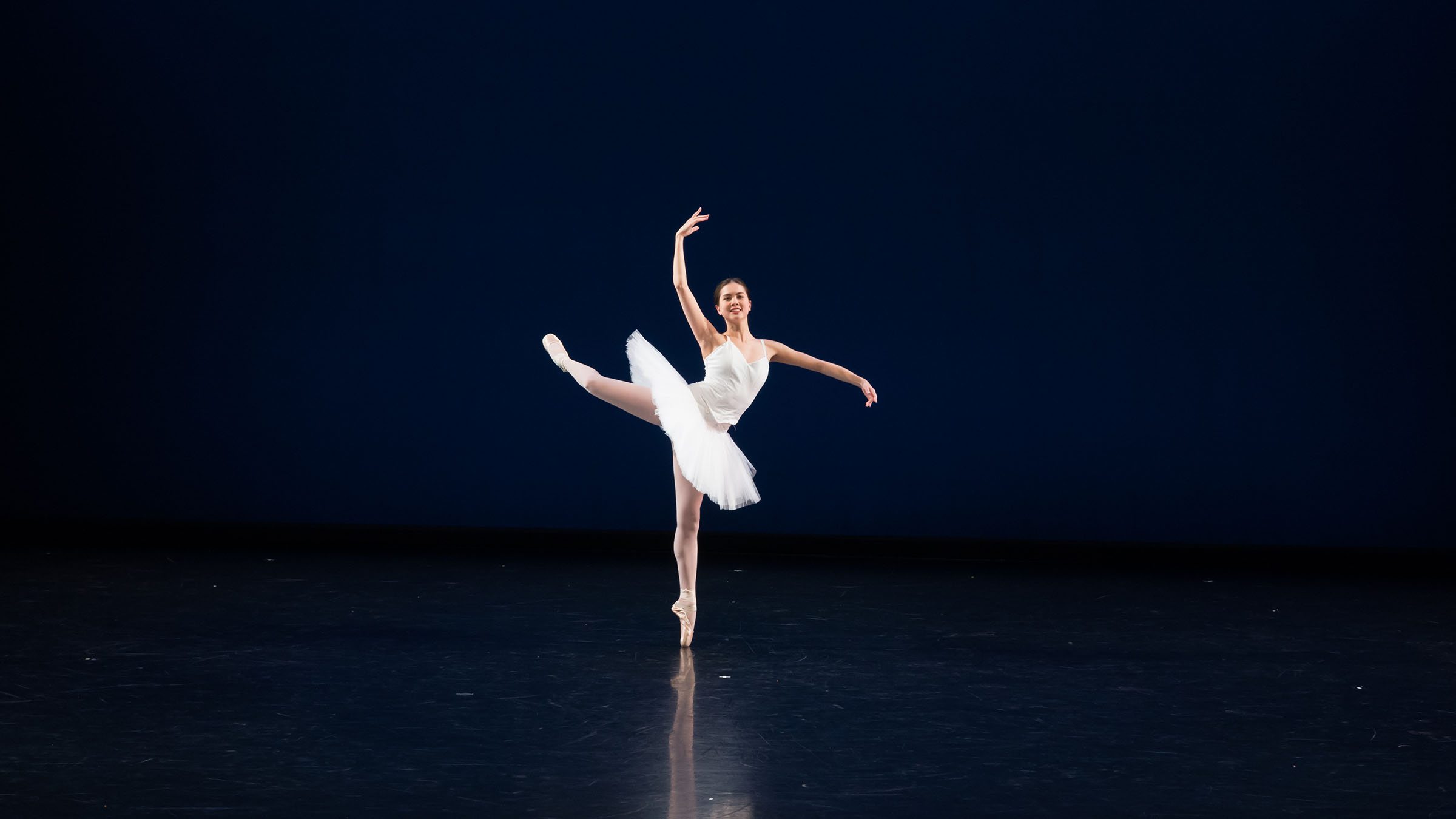 A person wearing a white ballet dress balances on one foot on a darkened stage. Photo by Mark Sugino.