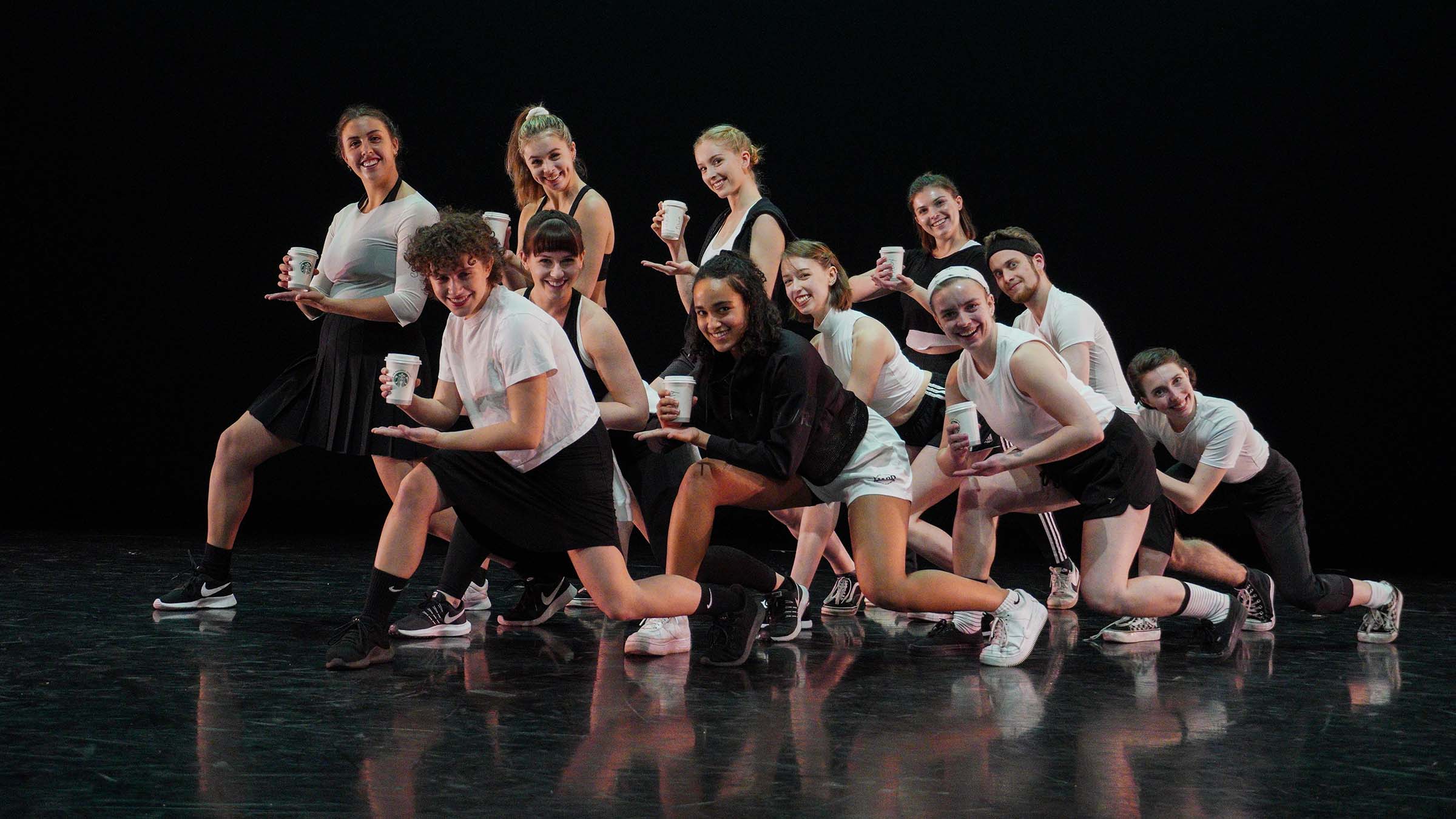 A group of dancers wearing casual clothing—mainly white shirts, black shorts, and sneakers—pose together while holding paper Starbucks coffee cups. Photo by Yesmina Townsley ’23