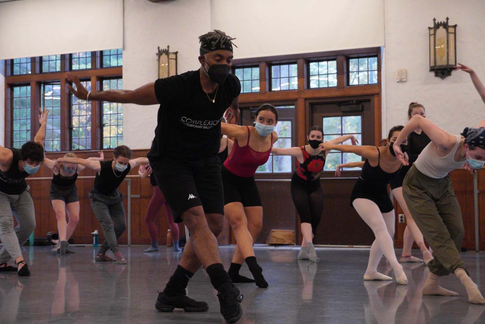 A person wearing a black T-shirt, black shorts, and a black facemask demonstrates a pose in a large, open classroom filled with other students who are also in that pose.