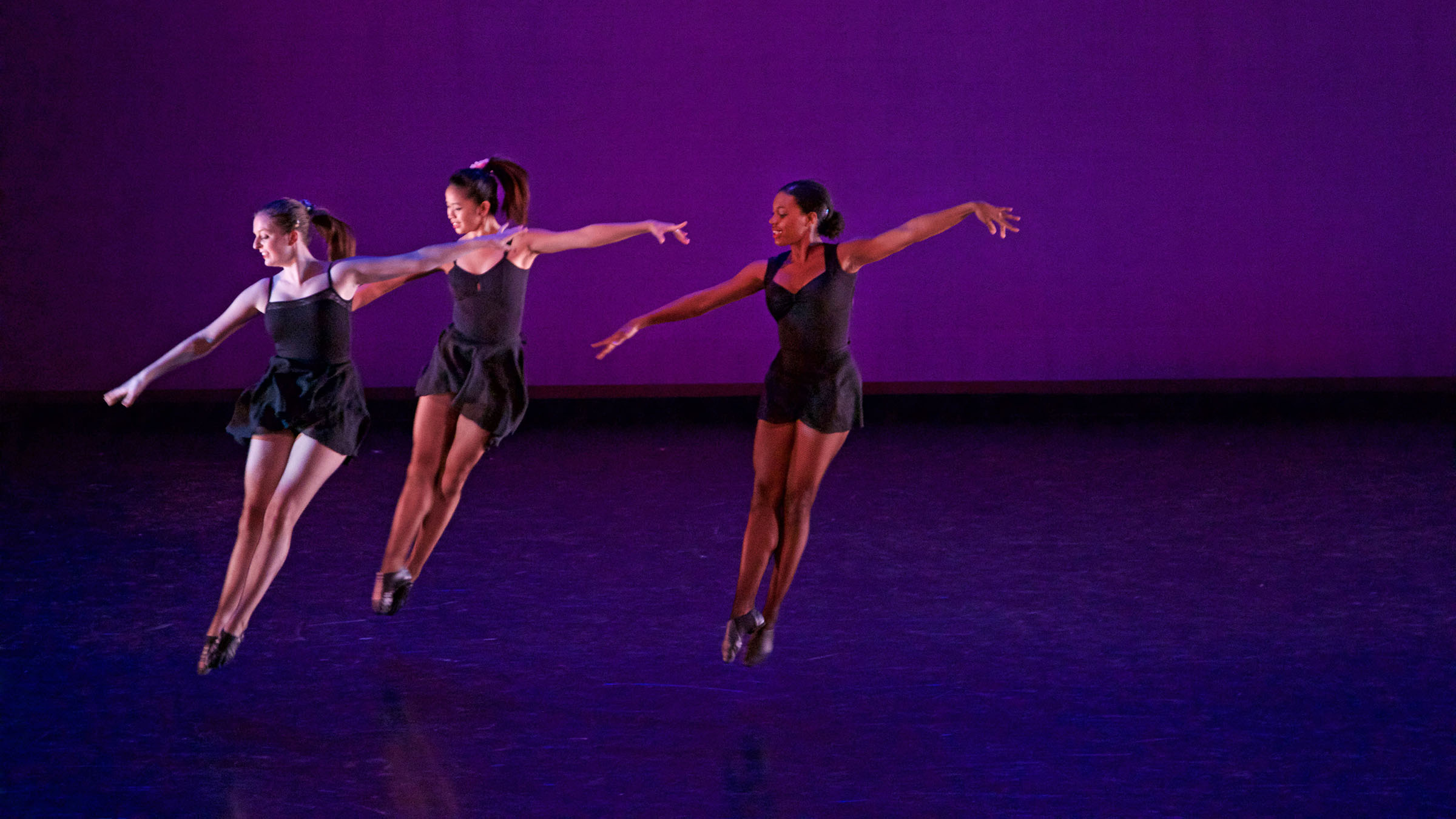 Three dancers leap in the air in unison on a dark stage. Photo by Mitchell Davis ’19.