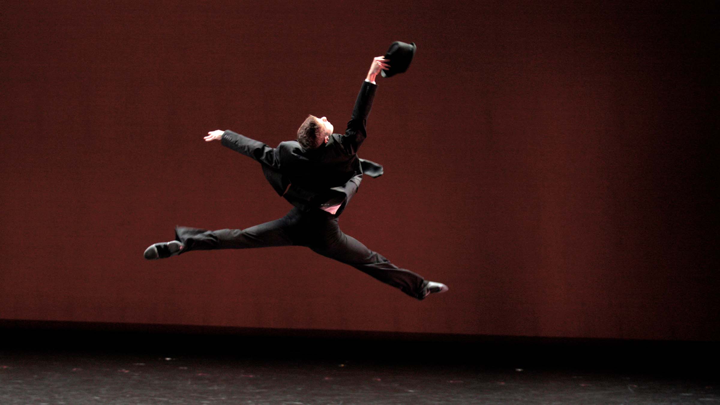 A dancer wearing a dark suit and holding what looks like a black fedora hat leaps across a stage. Photo by Rachel Garbade ’15.