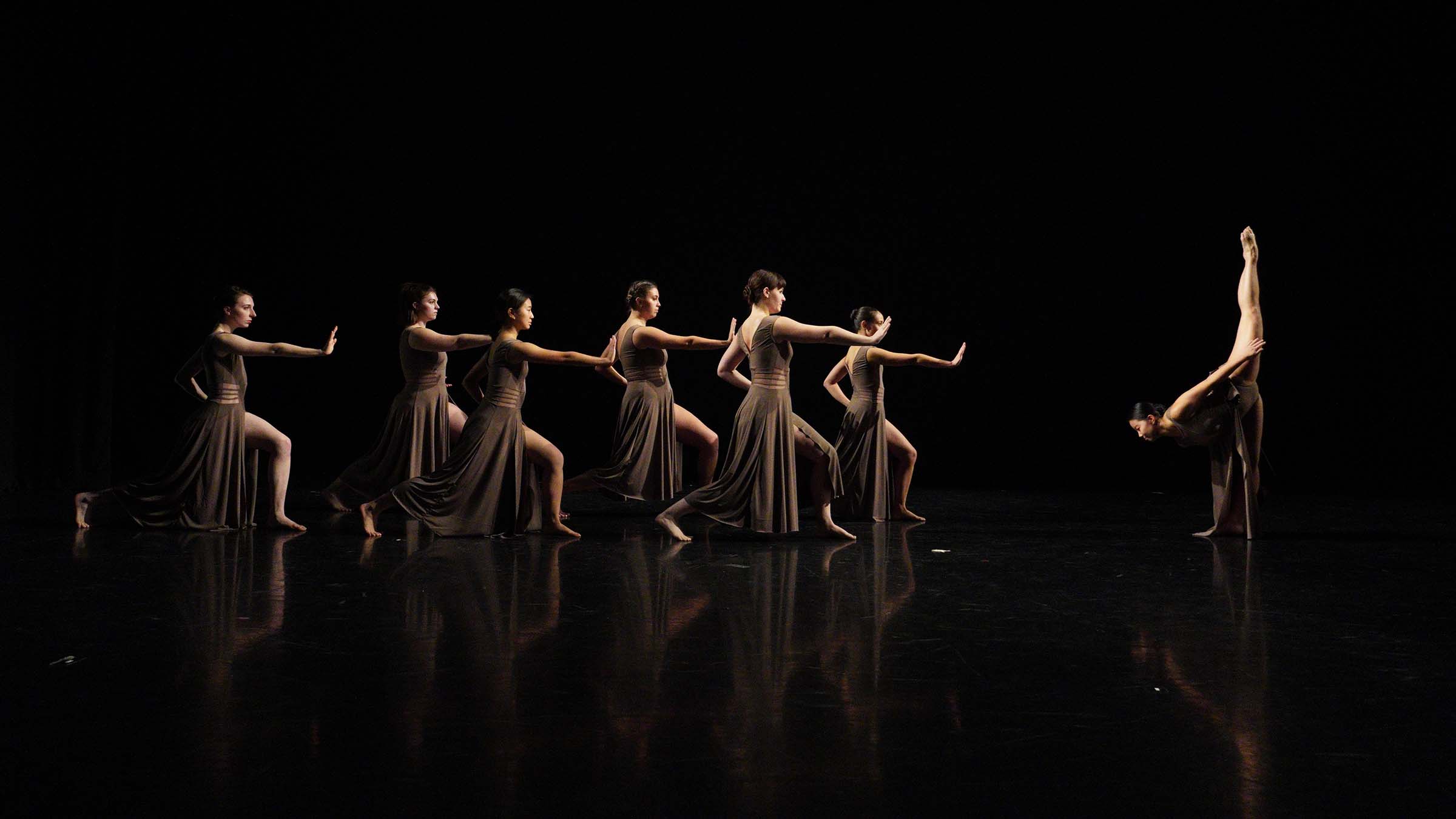 A group of dancers wearing long, brown dresses perform in synchrony on a darkened stage. Photo by Yesmina Townsley ’23.