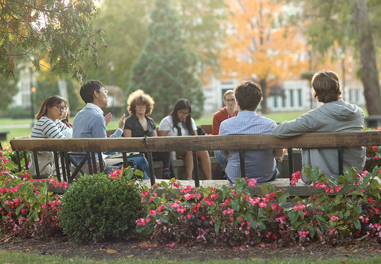 A group of students and a professor sit outdoors on a large circular wooden bench with flowering shrubs behind the bench.