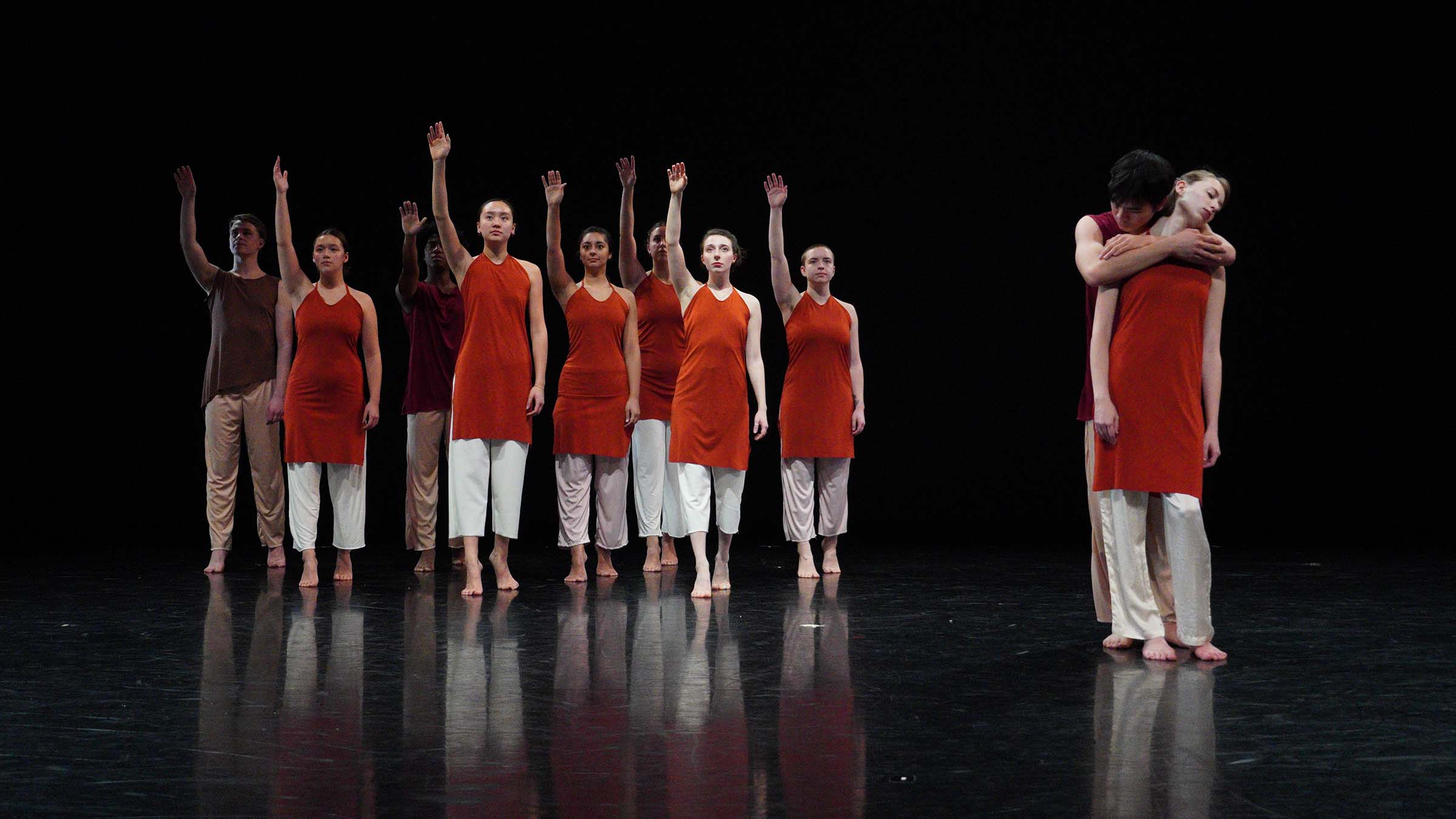 A group of people wearing white pants and long, red shirts, stand on a darkened stage with their right arms raised. To the right, two people stand alone; one person has their arms around the other person. Photo by Yesmina Townsley ’23
