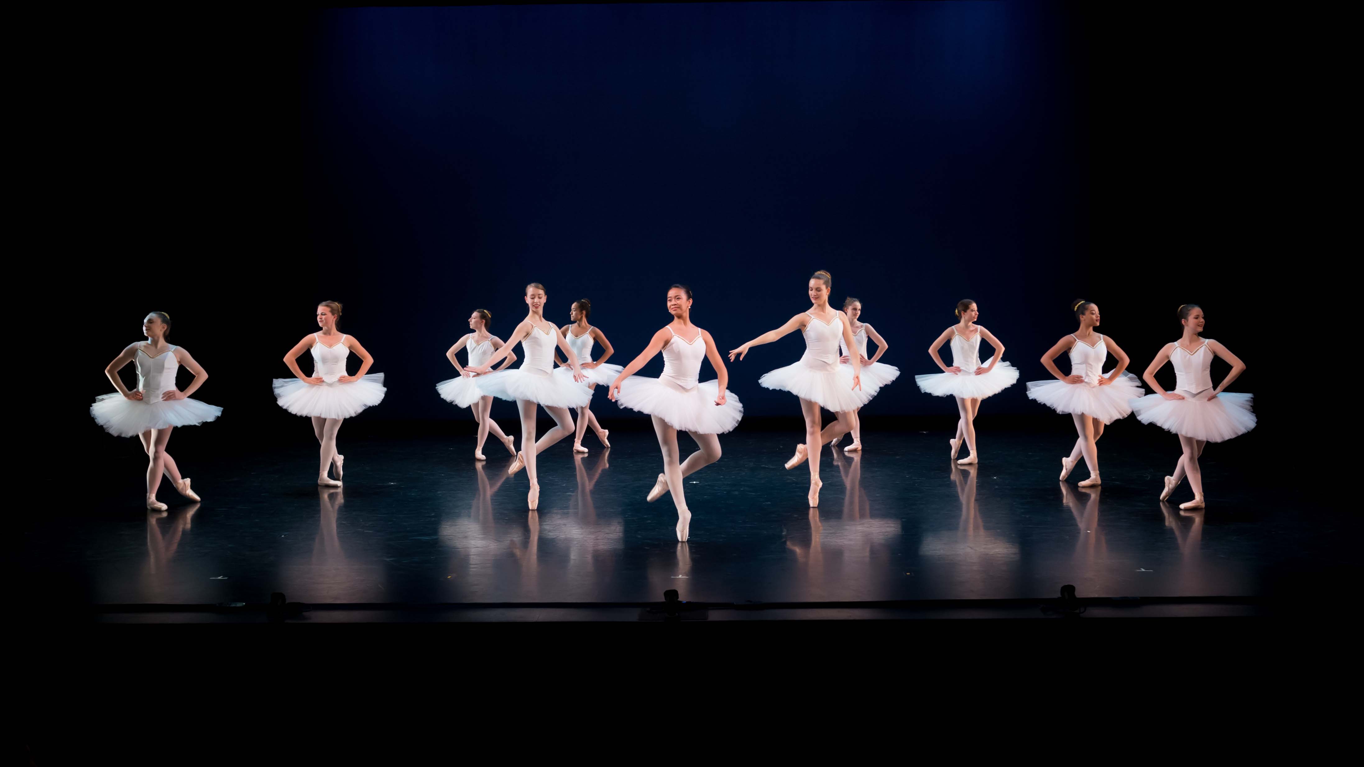A group of dancers wearing white ballet dresses perform in synchrony on a darkened stage. Photo by Mitchell Davis ’19.