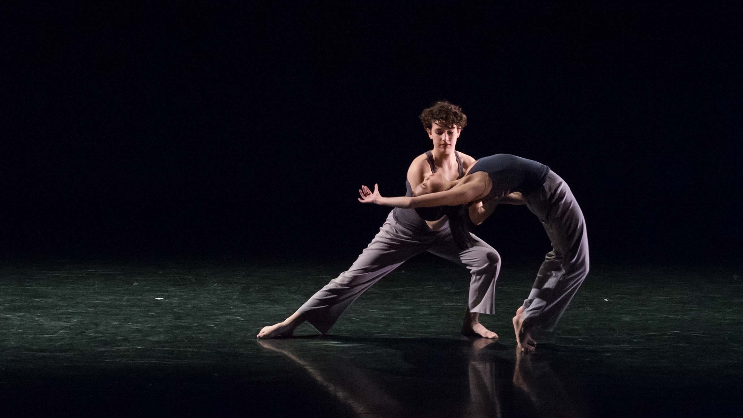 Two dancers perform acrobatics on a darkened stage. One dancer is bent over backwards, supported by the other. Photo by Mark Sugino.