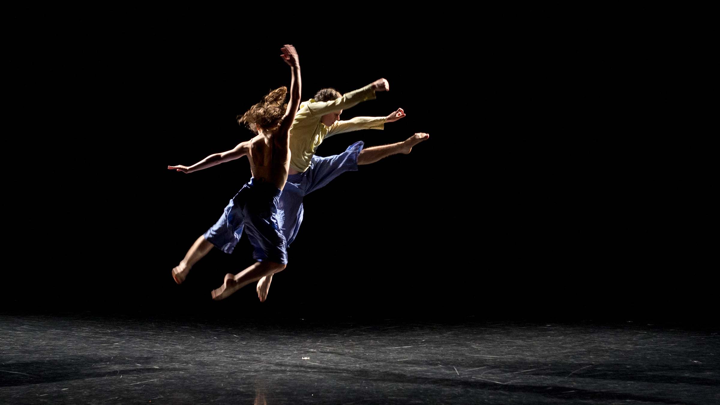 Two dancers caught in mid-leap on a darkened stage. Photo by Mark Sugino.