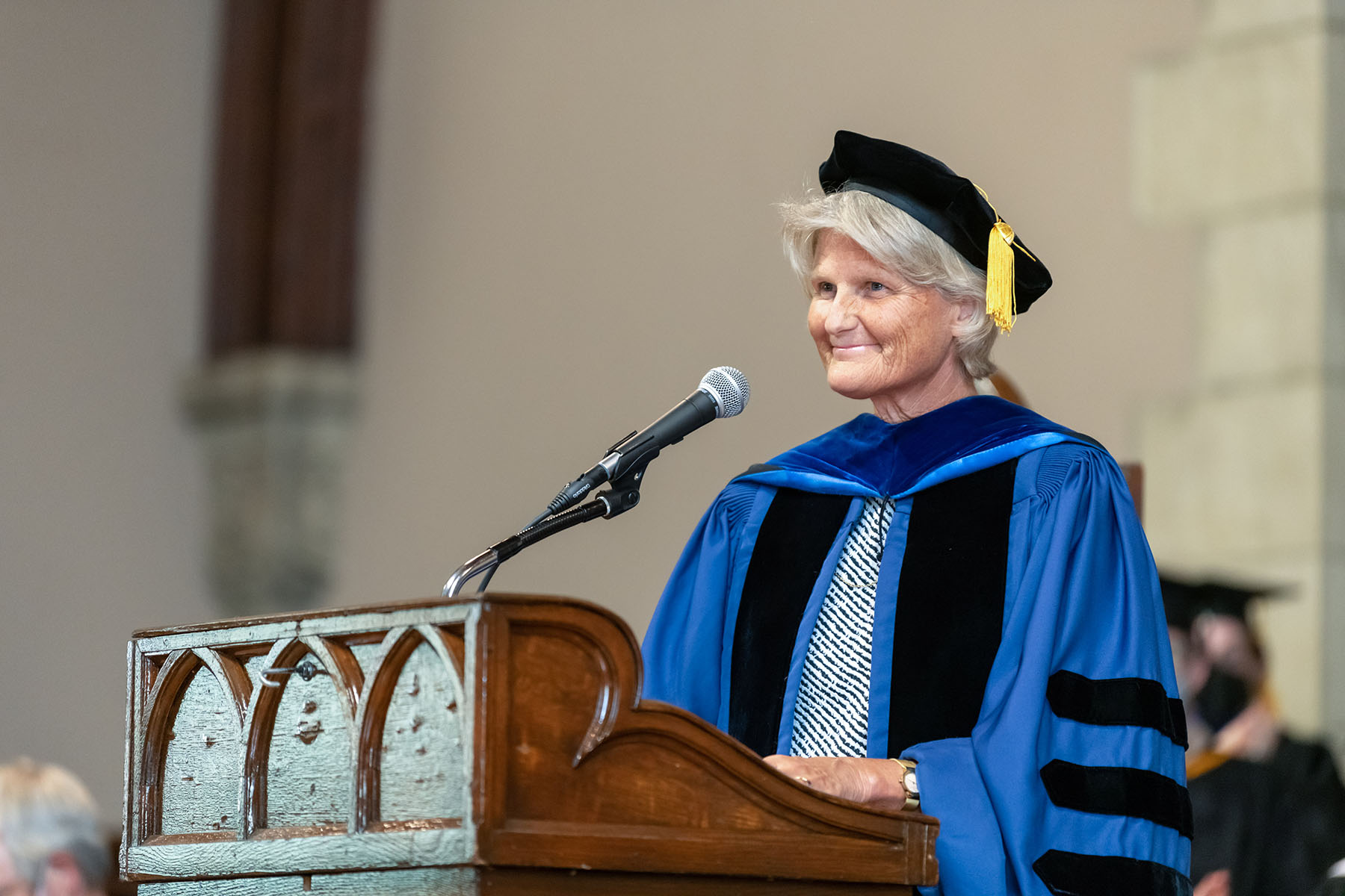 President Elizabeth H. Bradley speaks at a podium. President Bradley wears a blue robe and a black hat with a gold tassel.