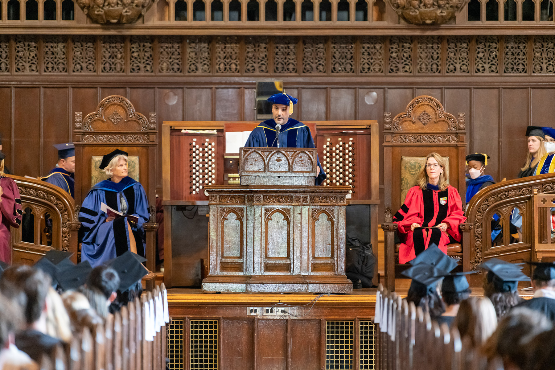 Dean of the College Carlos Alamo-Pastrana speaks at an ornate wooden podium, with members of the Vassar administration and faculty on both sides.
