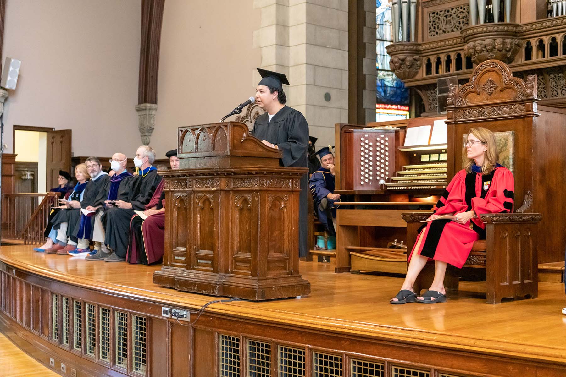 Julián Aguilar ’23, President of the Vassar Student Association, speaks on a large, wooden stage, with members of the administration and faculty on both sides. Aguilar is wearing a dark robe and dark graduation cap.