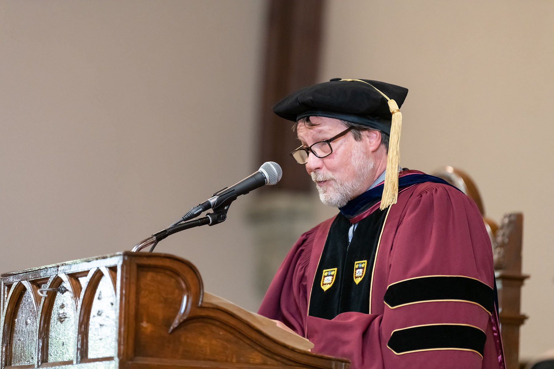 William Hoynes, Dean of the Faculty, speaks at a podium. Hoynes is wearing a burgundy robe and a dark cap with a gold tassel.