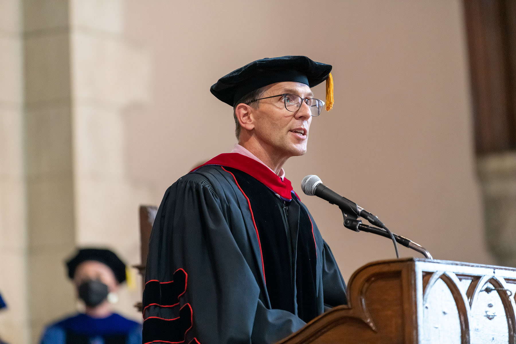 Reverend Samuel H. Speers stands at a podium, speaking. Reverend Speers is wearing a dark robe and a dark hat with a gold tassel.