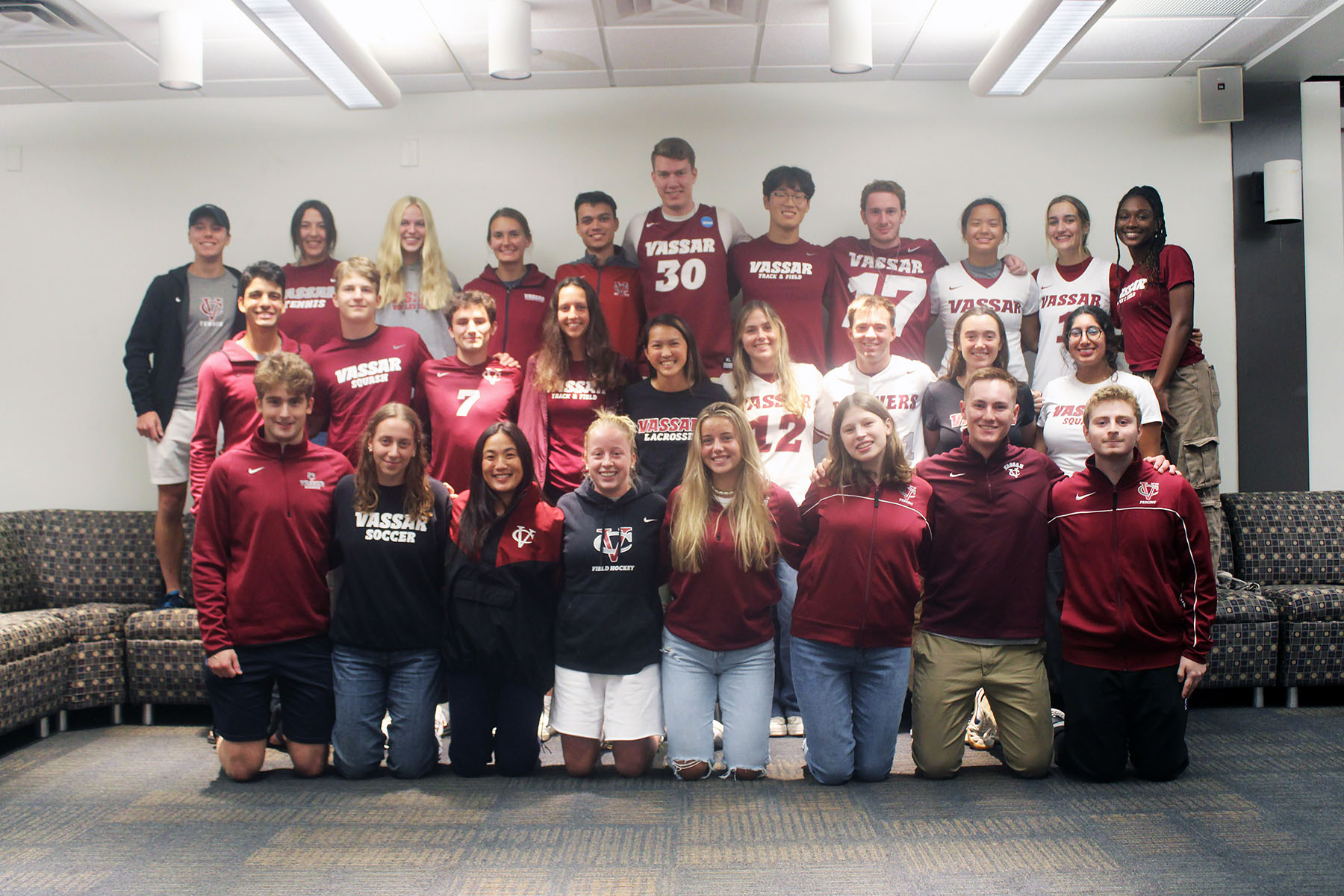 Group photo of the Student Athlete Advisory Committee wearing Vassar shirts and jackets kneeling and standing in front and on a couch. 