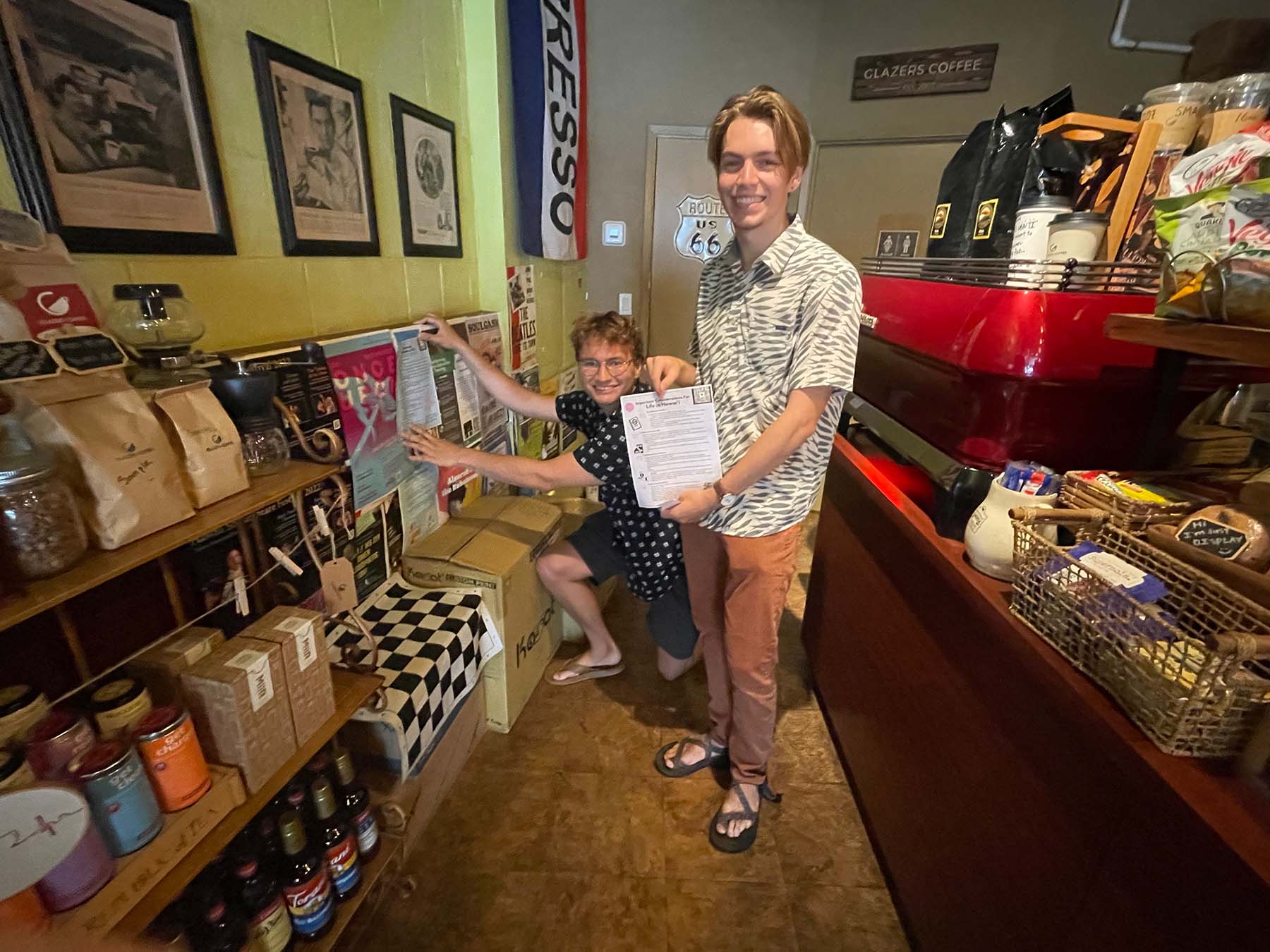 Christopher Unruh ’23 wearing navy blue patterned shirt, gray shorts and tan sandals and Chase Engel ’23 wearing gray and white patterned shirt, orange pants and brown sandals holding a document with immigrants’ legal rights inside Glazer’s Café next to shelves full of coffee products