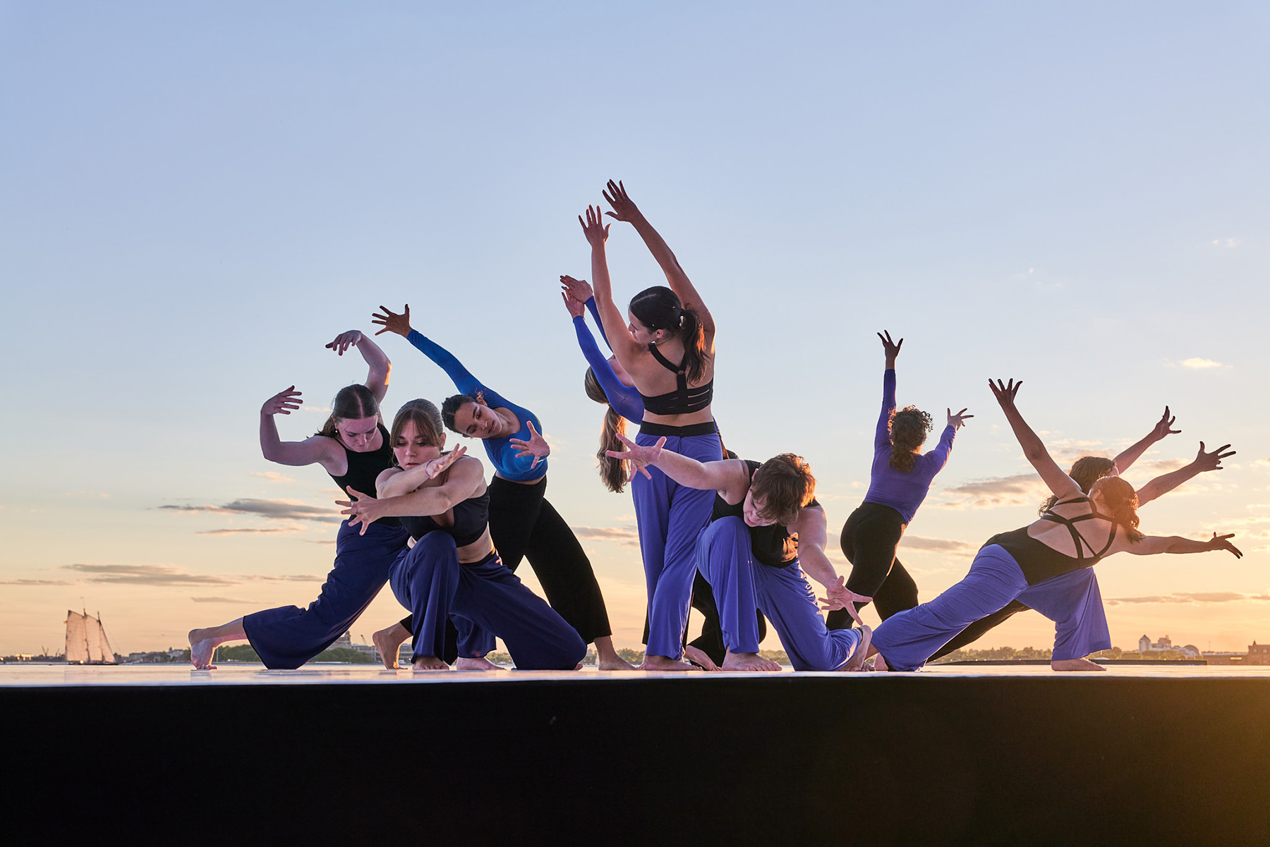 Dancers wearing black and blue in a pose with arms out and fingers spread with the sky in the background in a performance of Distance.
