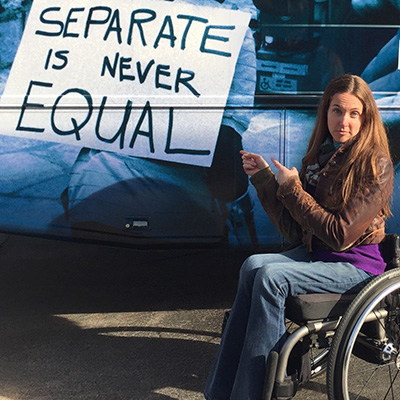 Disability advocate April Coughlin seated in her wheelchair and holding up a sign that reads "Separate Is never Equal"