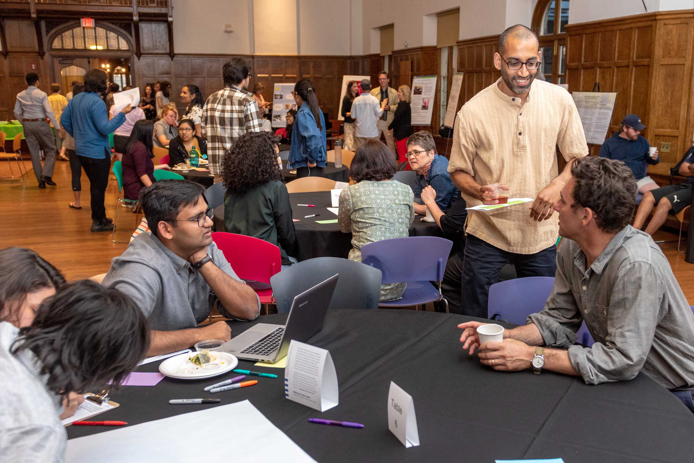 People sitting at a table and one standing in a crowded room discussing something. One person has a laptop