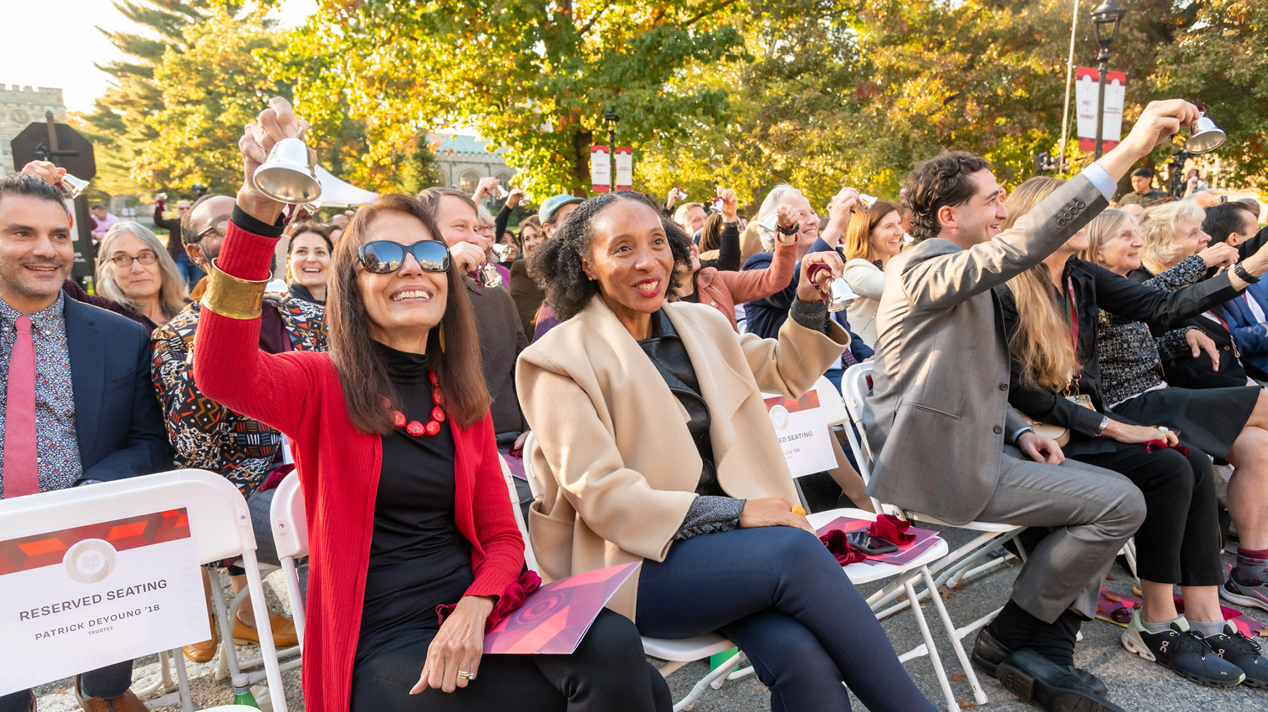 People sitting in an audience outside on folding chairs holding and ringing small silver bells