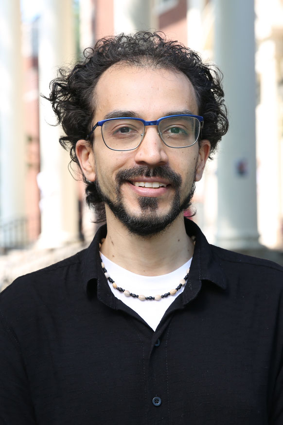A person with black, curly hair, a beard and mustache, glasses, and a black shirt smiles at the camera