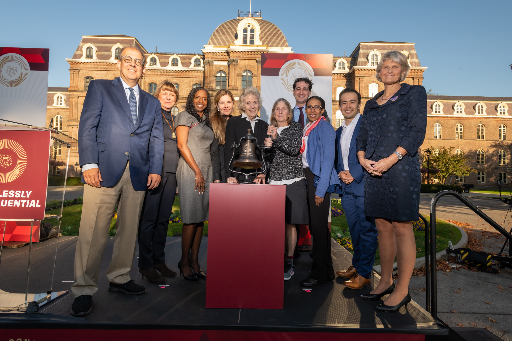 Ten people standing on a stage around the Vassar bell in front of Main building 