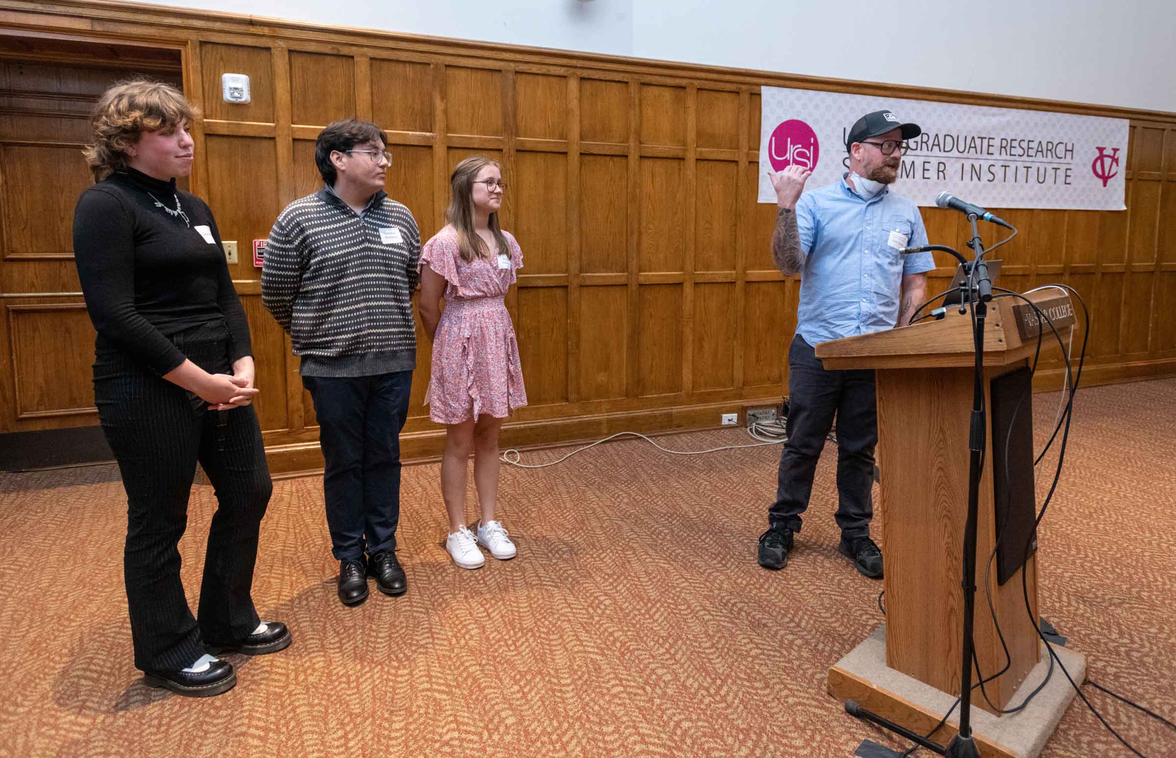 Three smiling students stand behind a podium facing an audience off screen while a person closer to the podium, also facing is audience, points at the students.