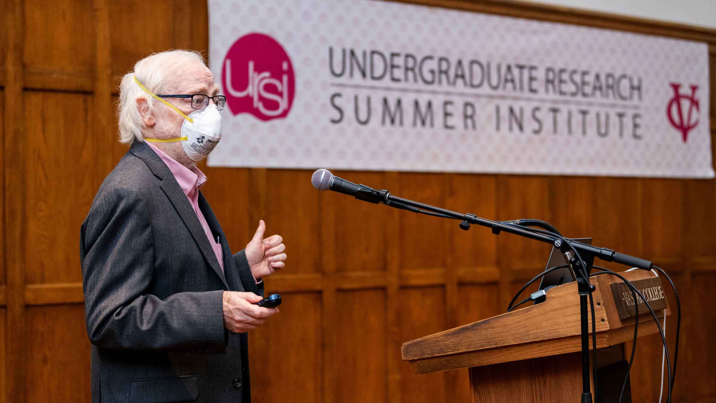 Masked speaker holding a slide clicker behind a podium with a microphone speaking to an audience off screen. There is a banner on the wall that reads: undergraduate research summer institute