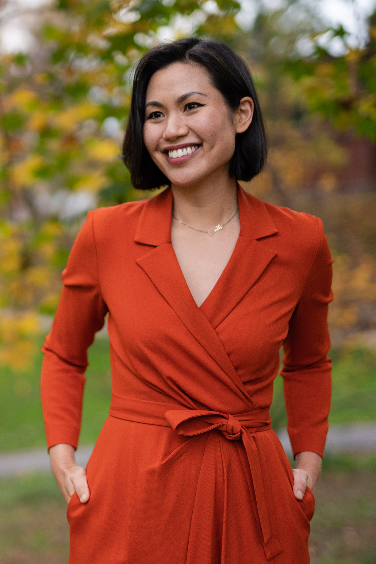 A person with long, black hair and a bright red-orange dress smiles at the camera