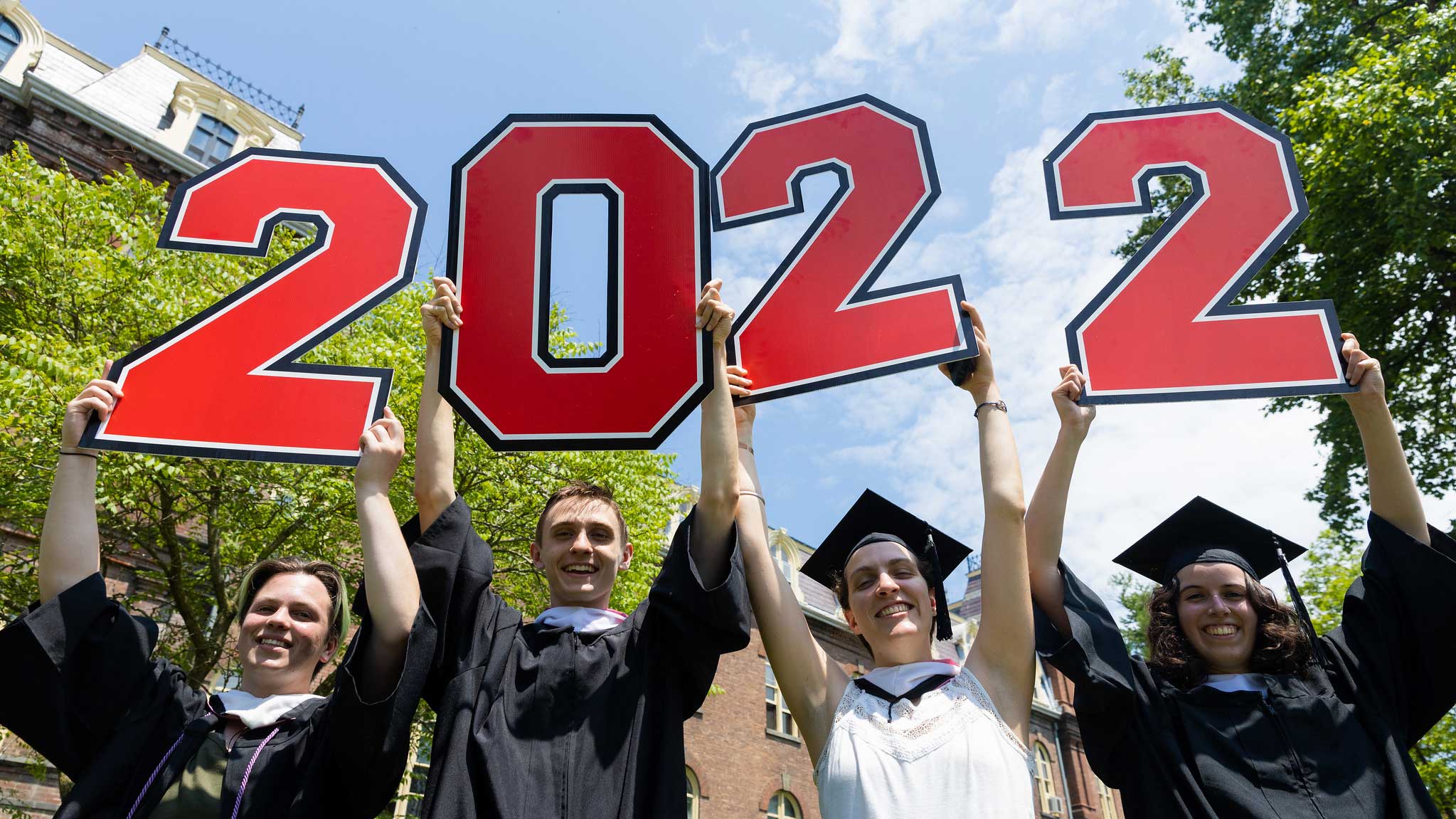 Four graduates in traditional garb standing in front of Vassar College Main Building holding up number signs that reads 2022