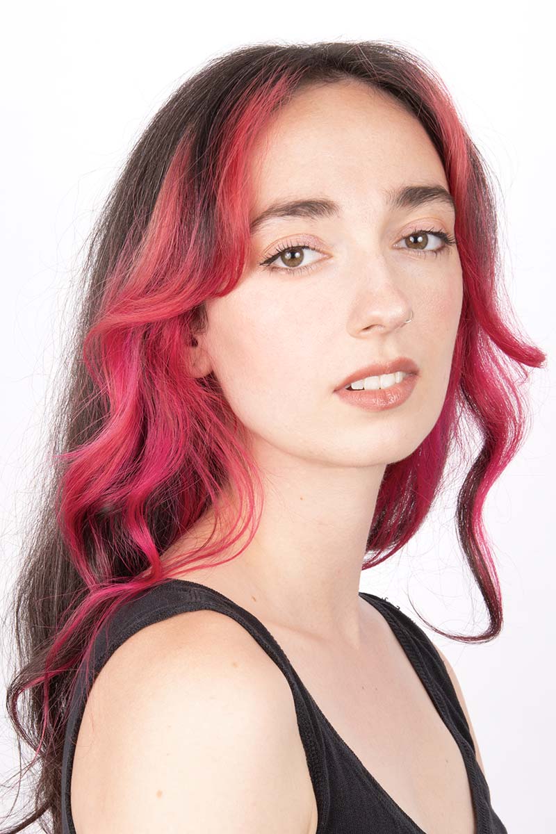 A photograph of a person with long, crimson hair wearing a black tank top and looking at the camera