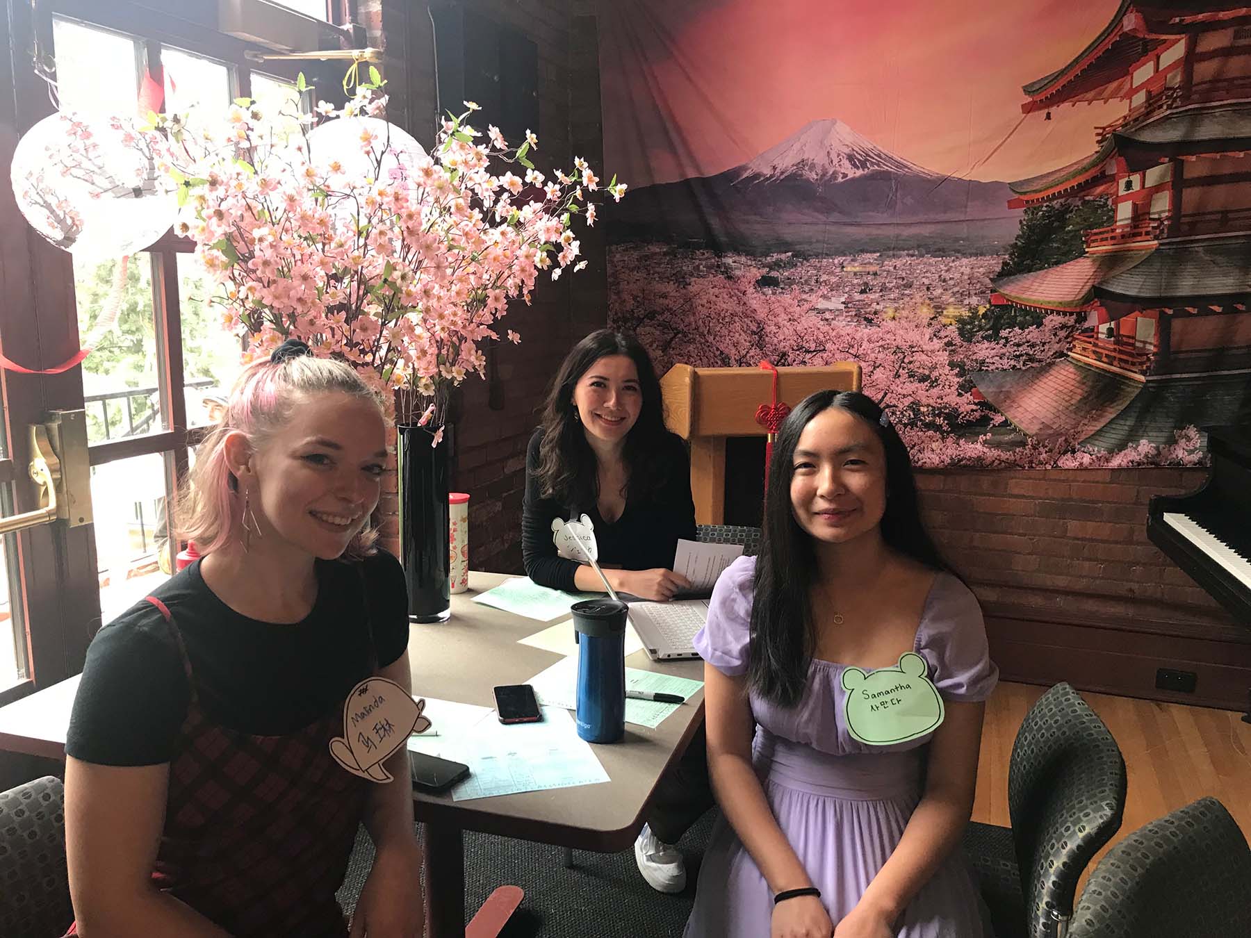 Three people sitting at a table for Chinese/Japanese Culture Day with a banner of Mt. Fuji and a Japanese temple in the background