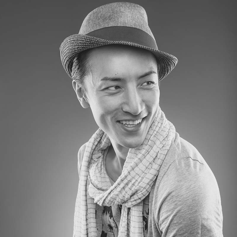 A grayscale photo of a person wearing a fedora hat and scarf, and smiling off-camera