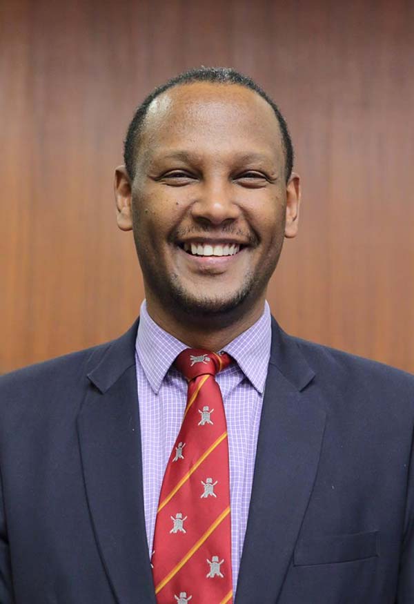 Dr. Abebe Bekele, a person with short dark hair and wearing a dark jacket with a red tie smiles at the camera