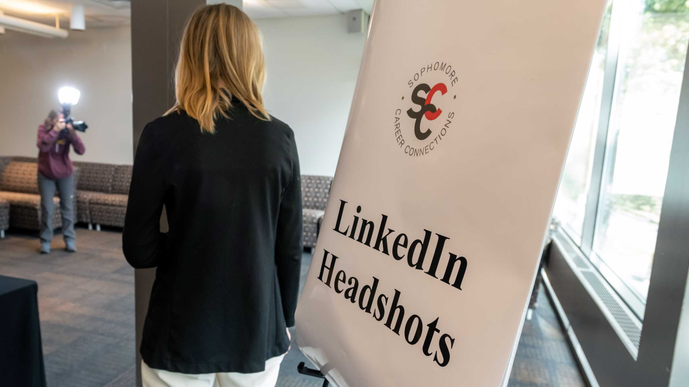 Person standing next to a sign the reads, LinkedIn Headshots as a photographer in the background snaps a flash photo of a person off camera.