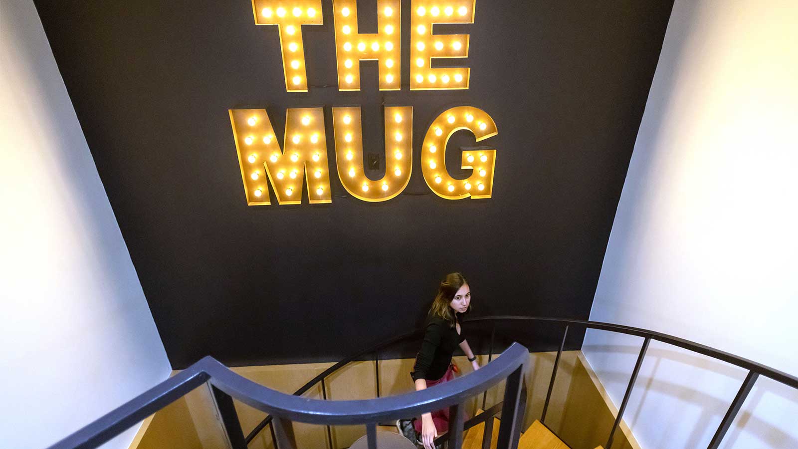 A person with long brown hair walks up a spiral staircase, underneath giant letters with lightbulbs that spell "The Mug."