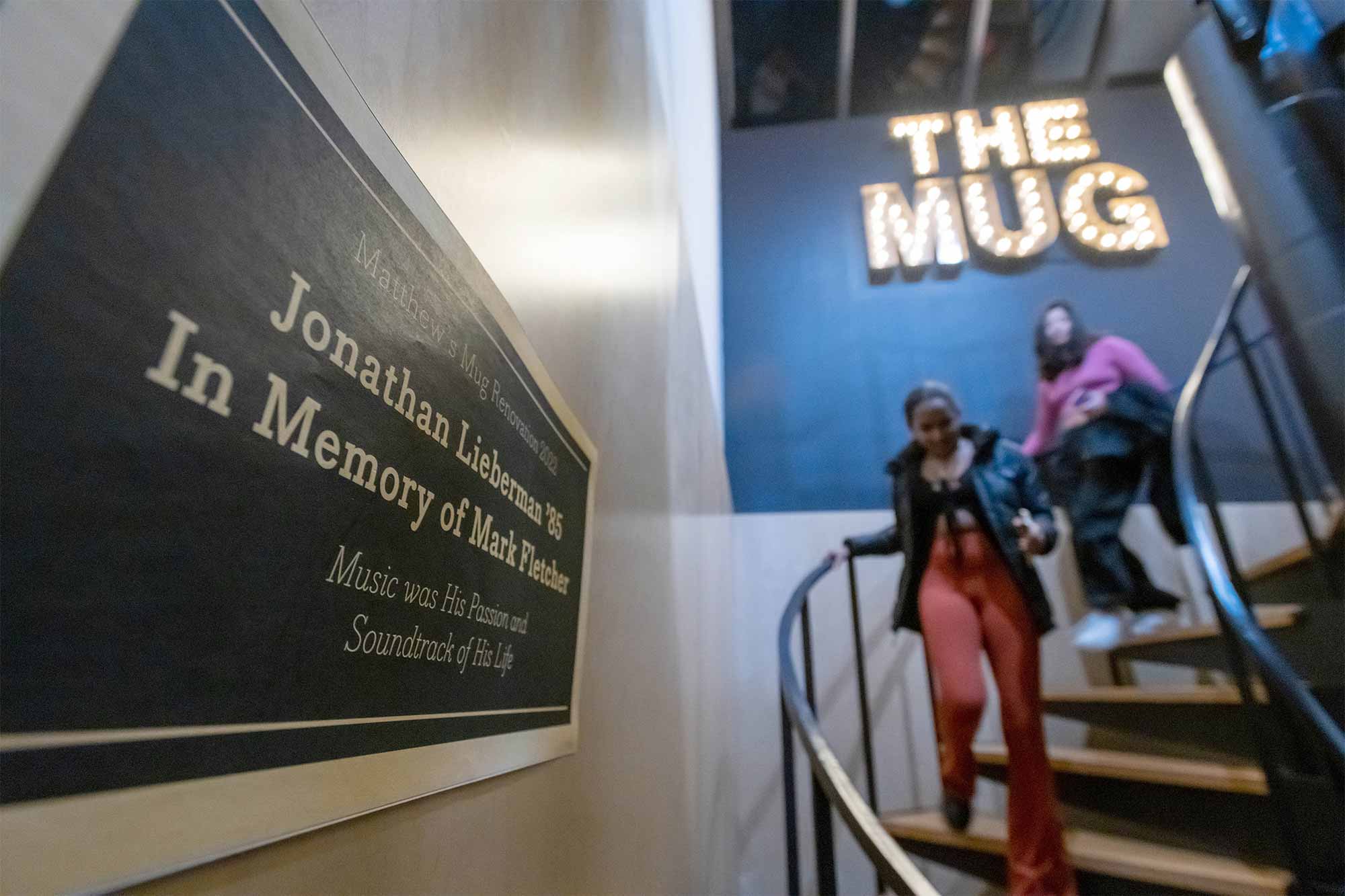 A closeup of a plaque that reads "Matthew's Mug Renovation 2022. Jonathan Lieberman ’85, In Memory of Mark Fletcher. Music was His Passion and Sountrack of His Life". In the background, two students descend a staircase under giant illuminated letters that read "The Mug".