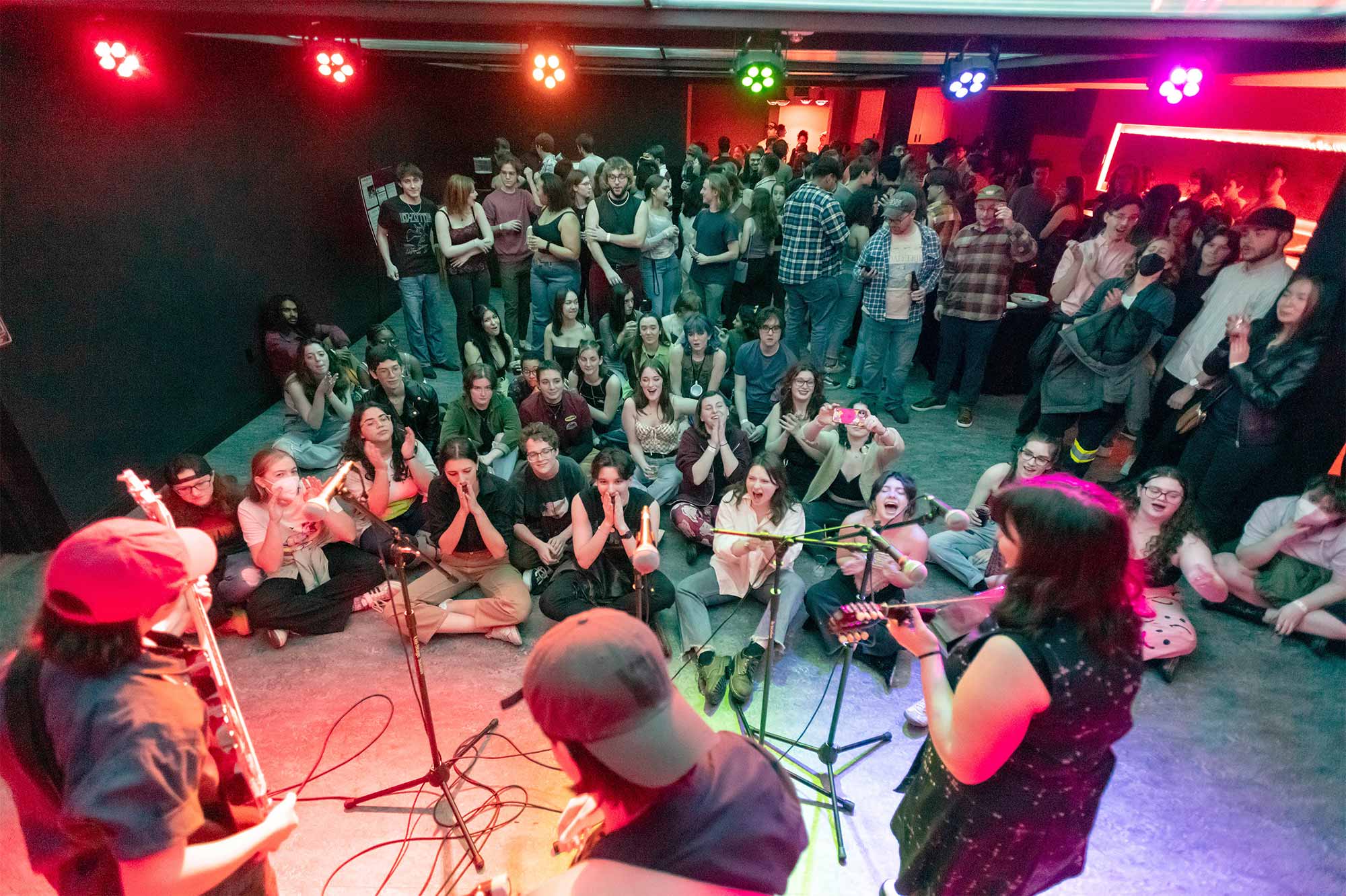 A room full of students. Many sit on the floor, watching a band play music. Colored lights hang overhead.