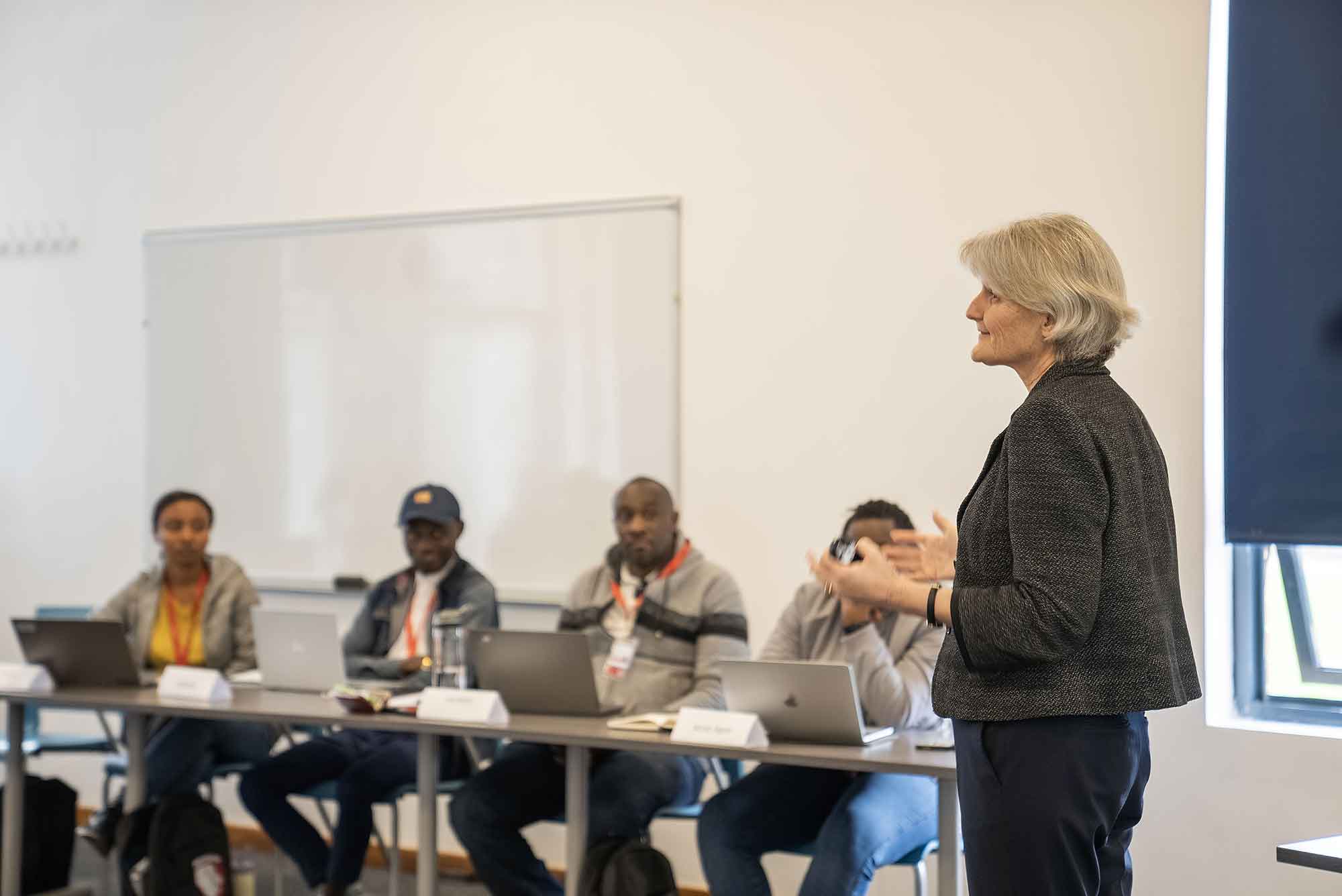 A classroom scene. Four people sit in front of laptops placed along a long table, listening to President Bradley speak. President Bradley is a person with shoulder-length gray hair and a dark gray shirt.