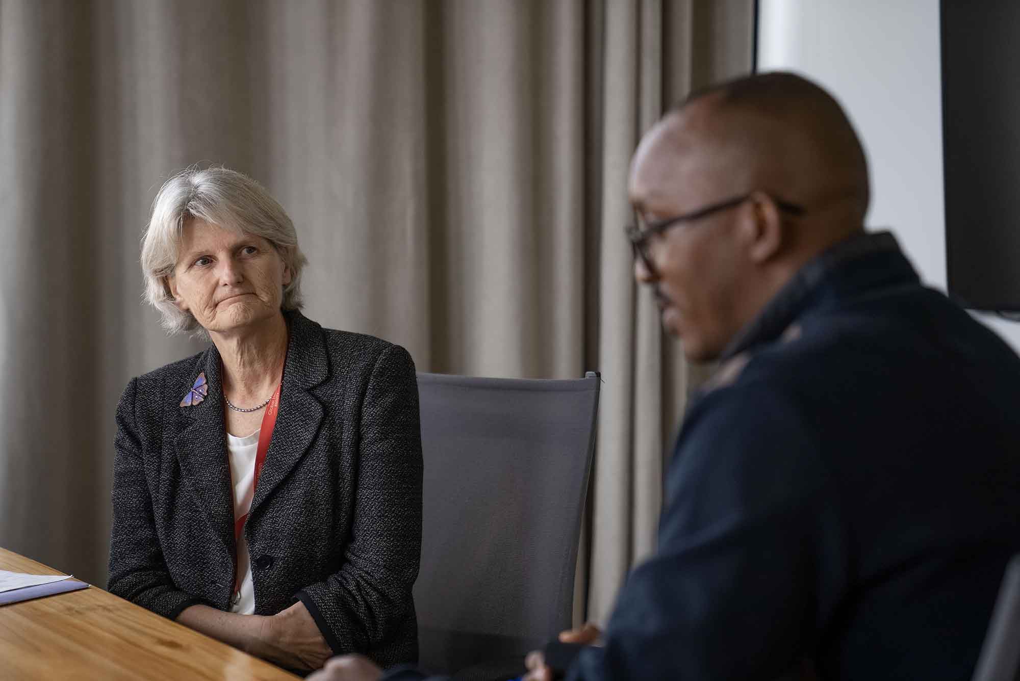 A closeup photo. President Bradley, a person with shoulder-length gray hair and a dark gray coat, sits at a wood-paneled table listening to a person with very short black hair, a thin black mustache, black eyeglasses, and a dark coat.