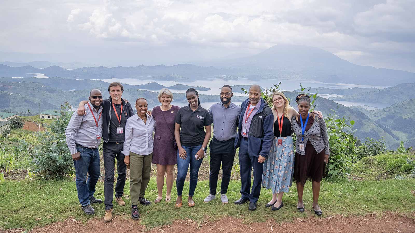 Nine people stand in a row on a grassy hilltop, smiling at the camera. Behind them is a view out over mountains and a river. The sky is filled with rolling clouds.