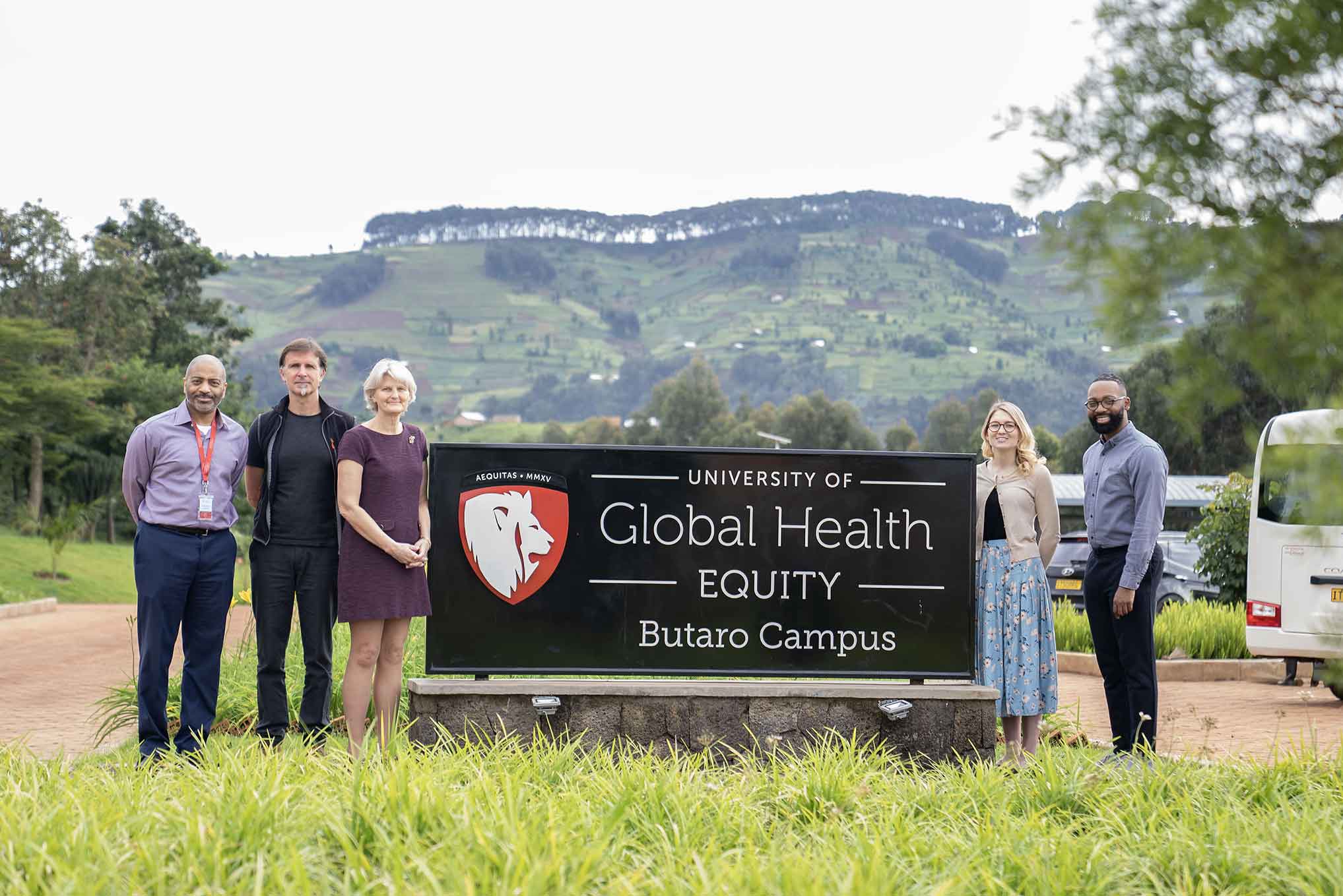 Five people stand next to a sign that reads “University of Global Health Equity; Butaro Campus”. The sign is placed in a grassy natural area with a farmland-covered hill in the background.