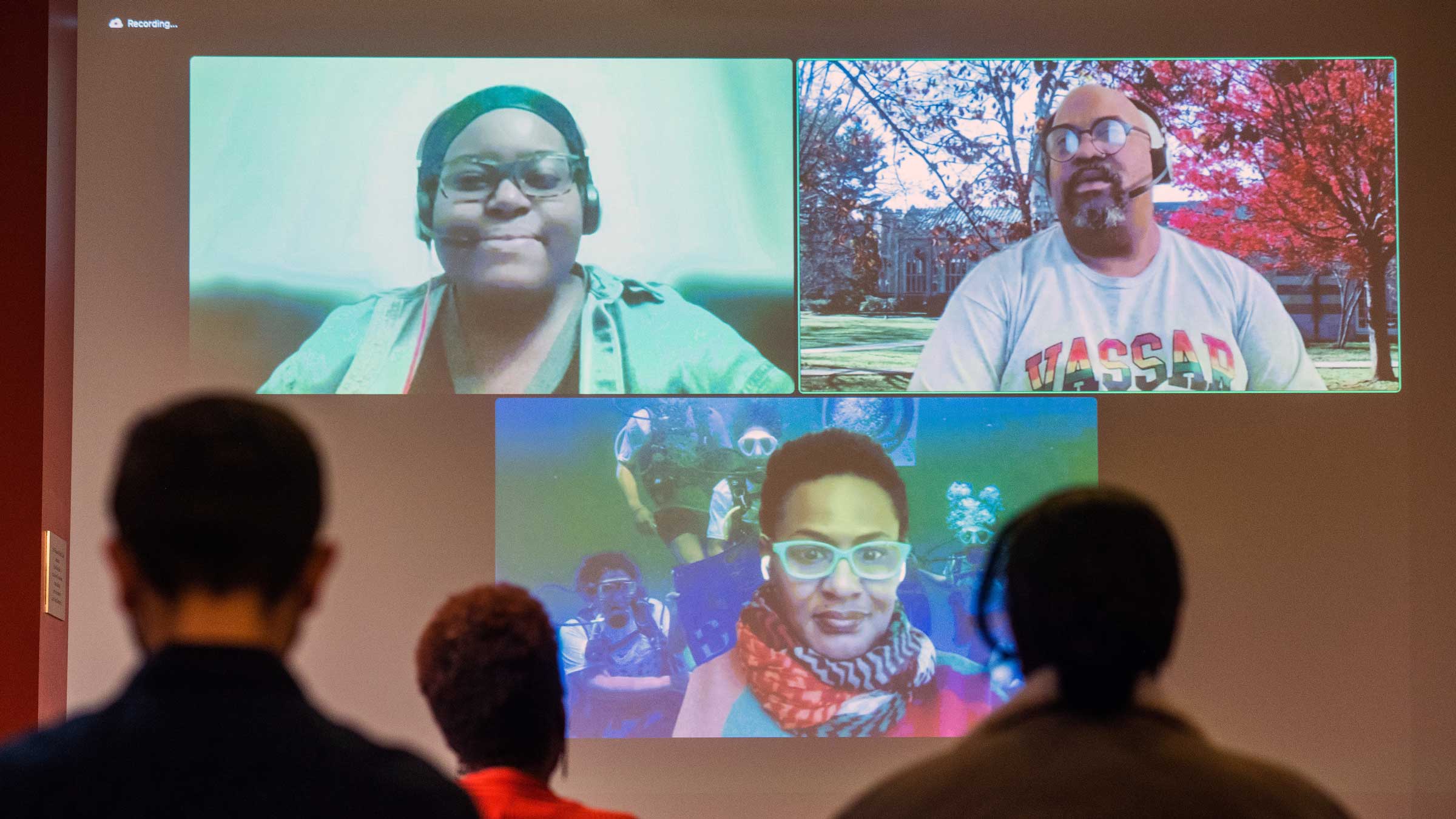 people with their backs to the camera seated in front of a screen with the projection of Zoom attendees in the same meeting