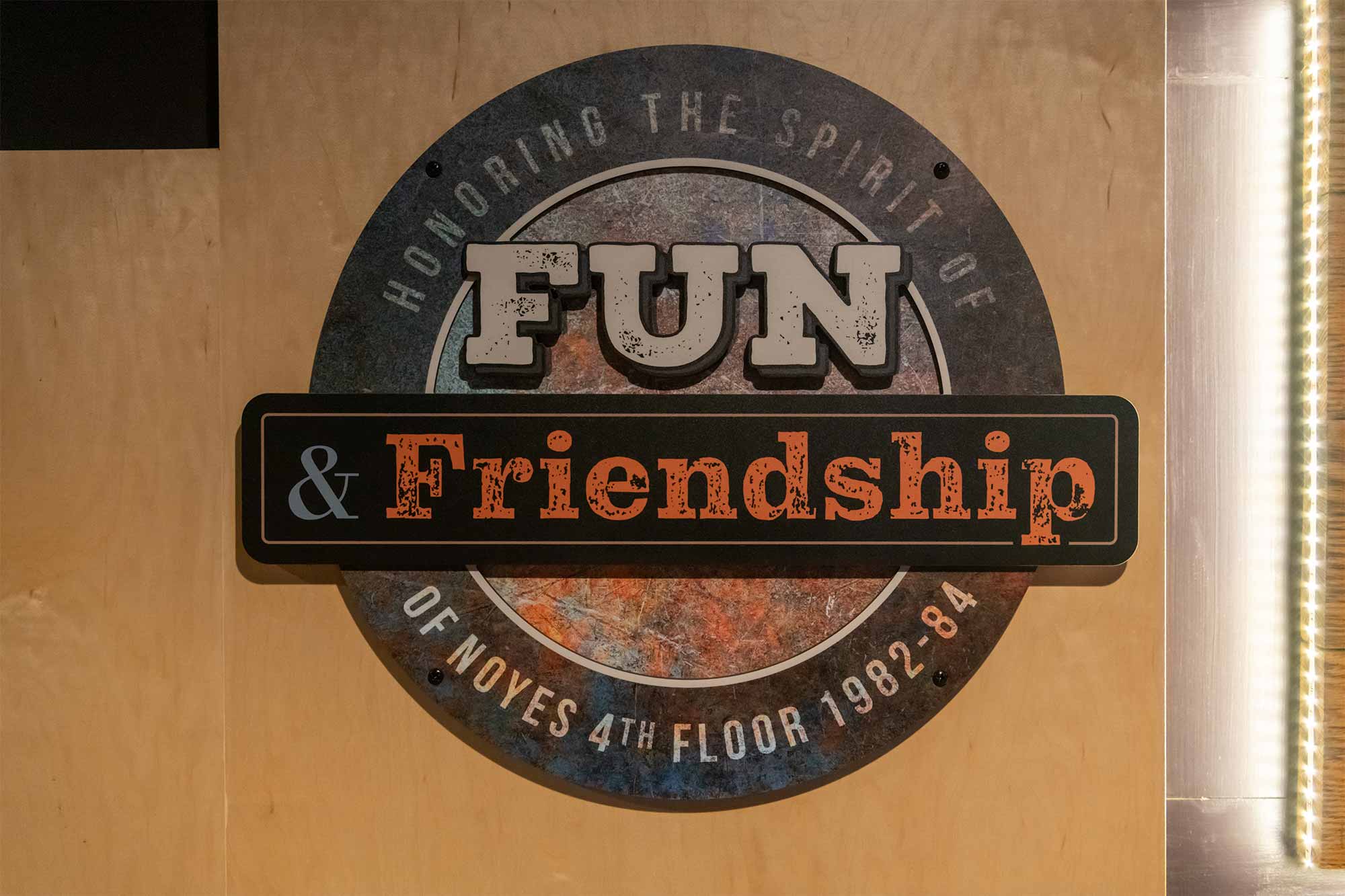 A round sign hung on a door. The sign has the text "Honoring the spirit of Fun and Friendship of Noyes 4th Floor 1982–84"