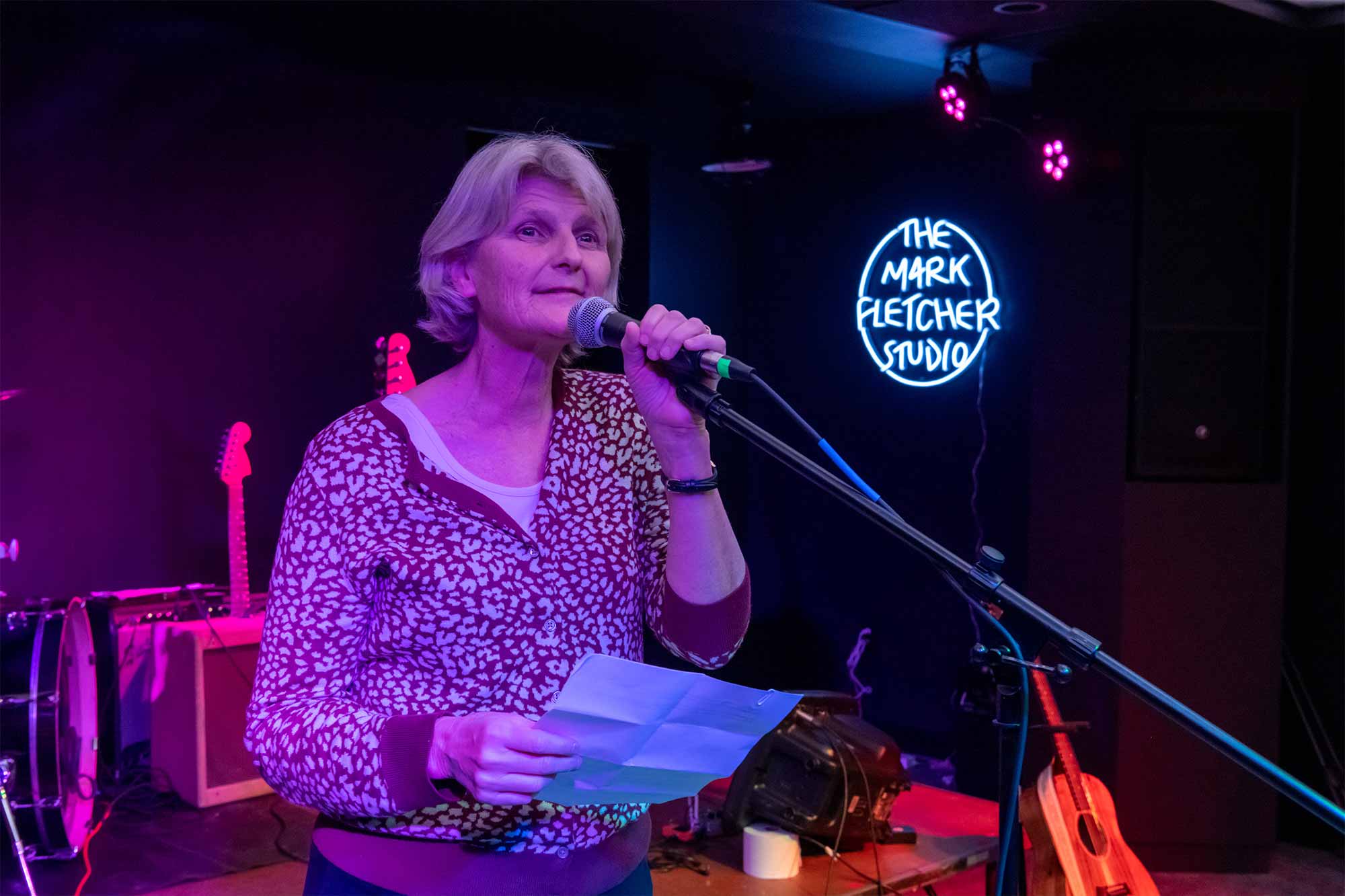 A person with mid-length gray hair stands in a dark room lit by neon colors, speaking into a microphone while holding a sheet of paper.