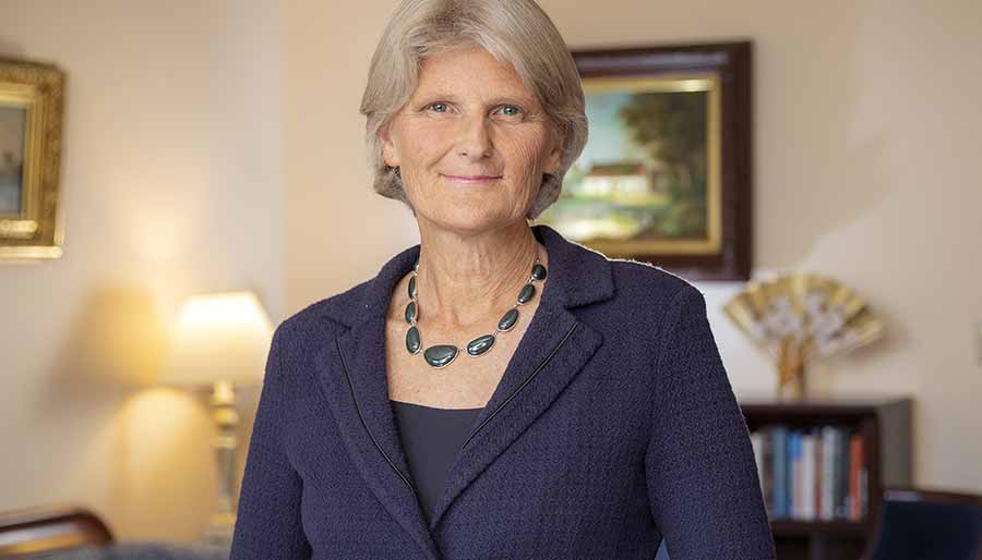 A person with straight gray hair, a necklace of shiny black stones, and an indigo jacket smiles at the camera