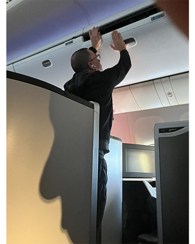Inside an airplane cabin, a passenger reaches up to stow luggage; a large shadow of a face covers a wall nearby.